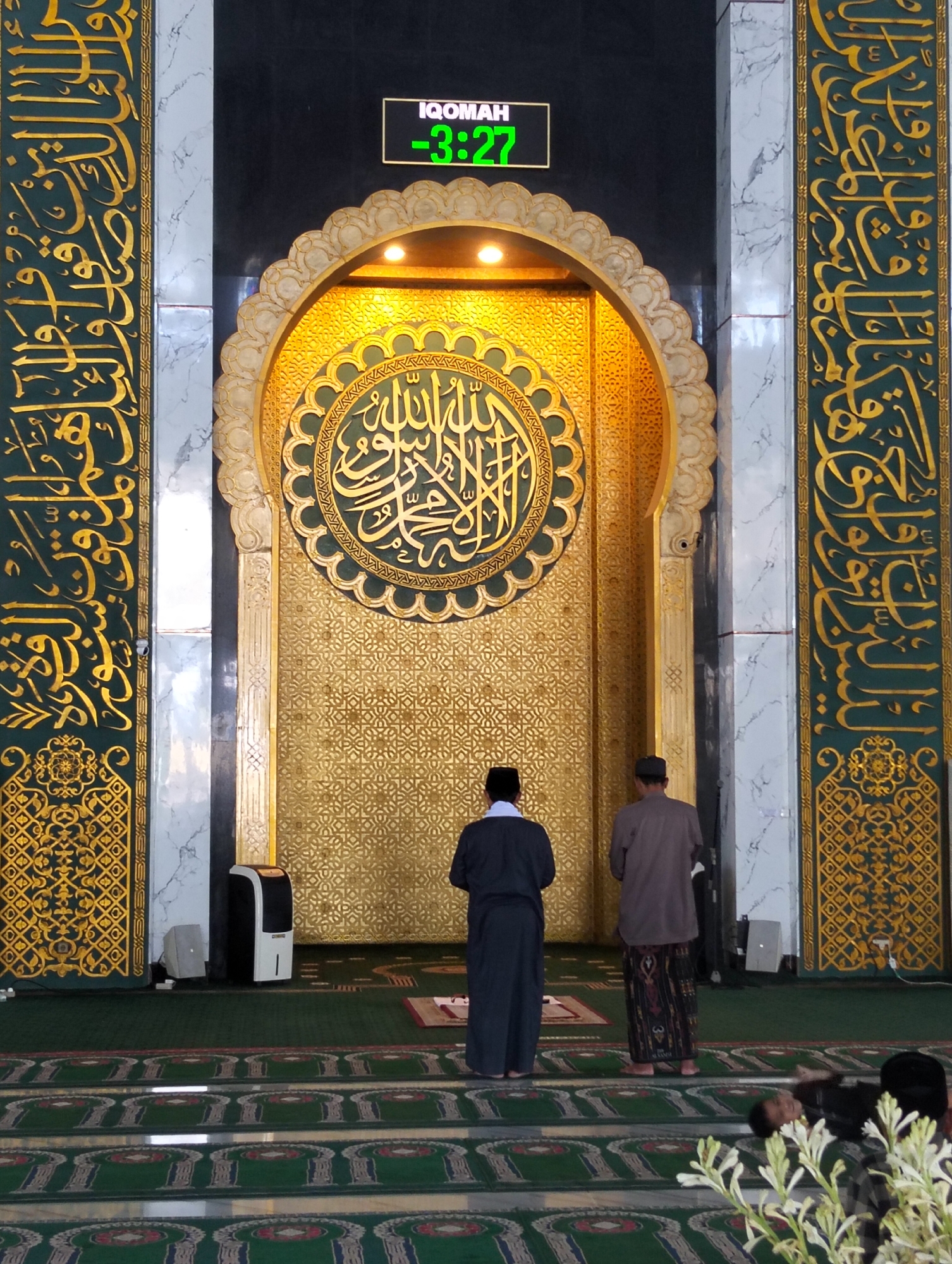 The image depicts the interior of a mosque, likely in Surabaya, Indonesia, with a focus on the mihrab area. Two men are standing in prayer facing the mihrab, which is adorned with intricate golden Islamic calligraphy and geometric patterns. Above the mihrab, a digital clock displays "IQOMAH -3:27", indicating the time remaining until the commencement of prayer. The walls of the mosque are decorated with ornate green and gold Islamic calligraphy. In the foreground, a green carpet with intricate patterns covers the floor, and a person is lying down on the carpet. There are also a few white flowers in the bottom right corner of the image. The overall impression is one of a sacred and tranquil space.