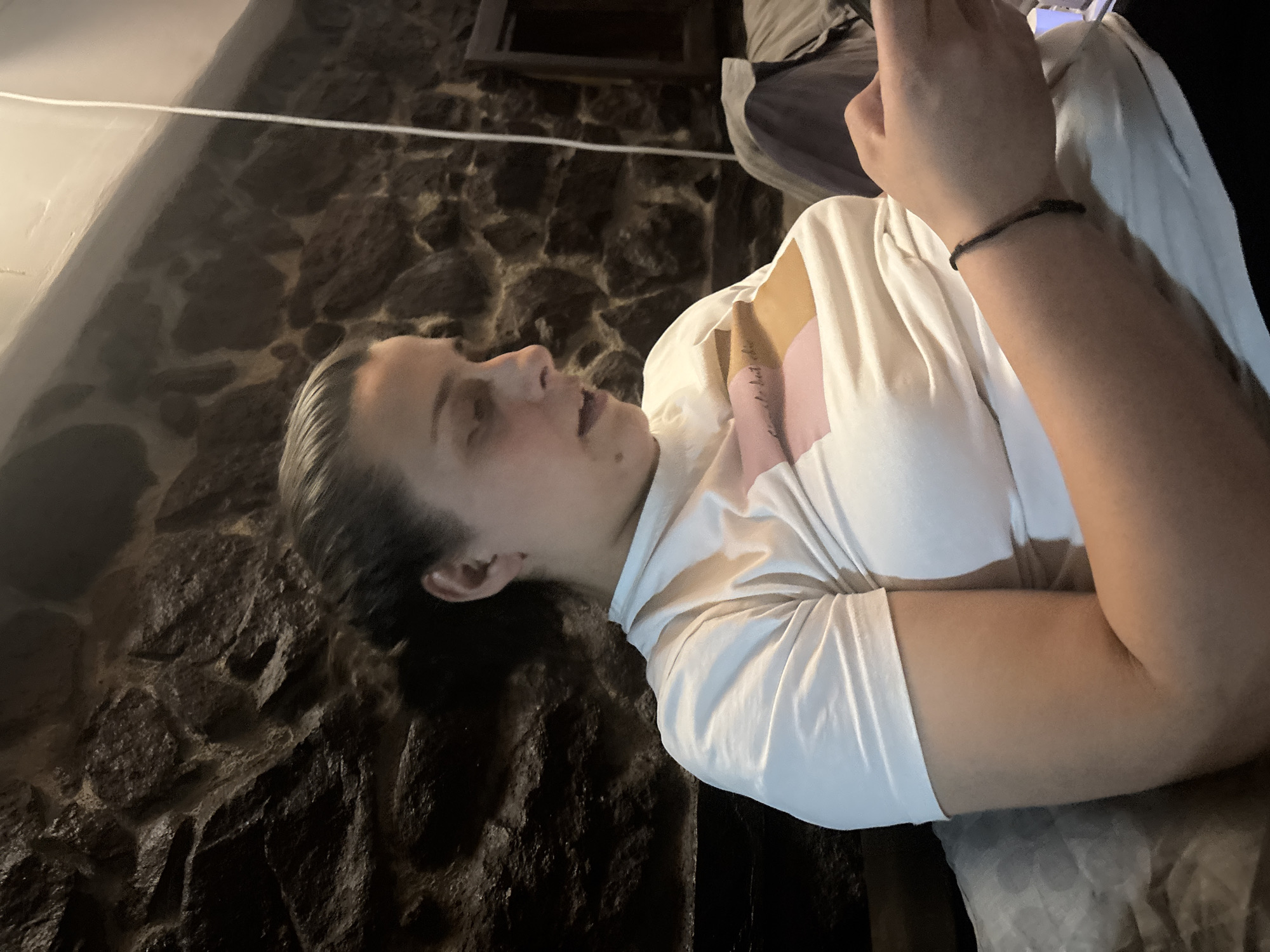 The image, rotated 90 degrees clockwise, features a young person lying on their side against a dark, textured stone or brick wall, typical of an old building in Belopoptsi, Bulgaria. The dim lighting suggests it is evening or night indoors.
The subject has dark, slicked-back hair and a neutral expression, looking upwards. They are wearing a white t-shirt, with a glimpse of a light pink garment or design visible beneath it on their right shoulder and chest. A black string bracelet is on their left wrist. Their hands are clasped, holding a smartphone, which is partially visible in the upper right.
Faint, indistinct text, possibly a tattoo like "Je te veux", can be seen on their right inner arm. A thin white wire or string crosses horizontally through the upper background. The stone wall provides a rustic backdrop, and a dark, rectangular shape (possibly a window or frame) is visible higher on the wall. The person's right arm rests on a light grey, textured surface, possibly a cushion or bedding. The activity appears to be the person relaxing and engaging with their phone.