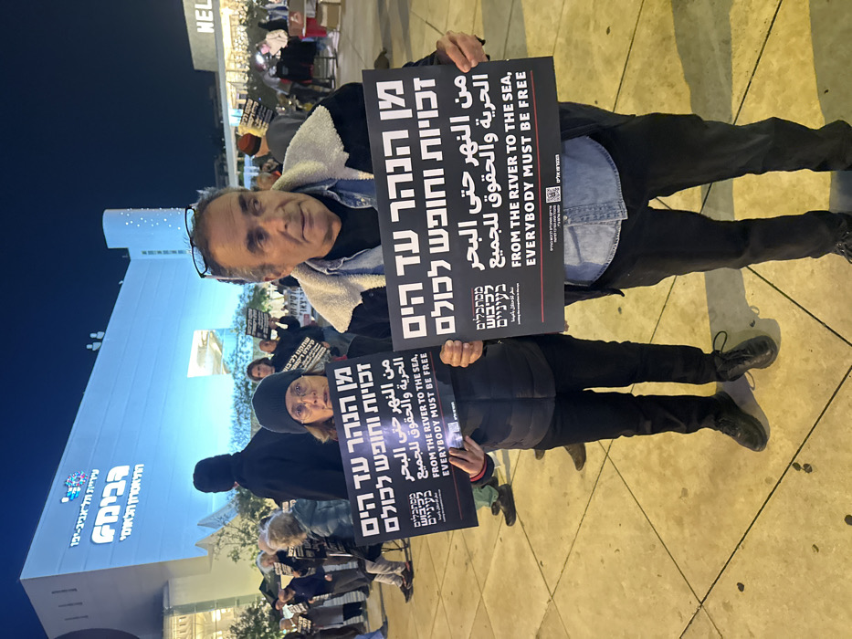 A group of protestors hold signs that read in Hebrew, Arabic, and English, "From the river to the sea, everybody must be free." The signs also include a QR code. The protestors appear to be standing outside a building labeled in Hebrew, "Tel Aviv-Yafo Municipality. Habima National Theatre." The sky is dark, suggesting it's evening. The scene appears to be a political demonstration or rally.