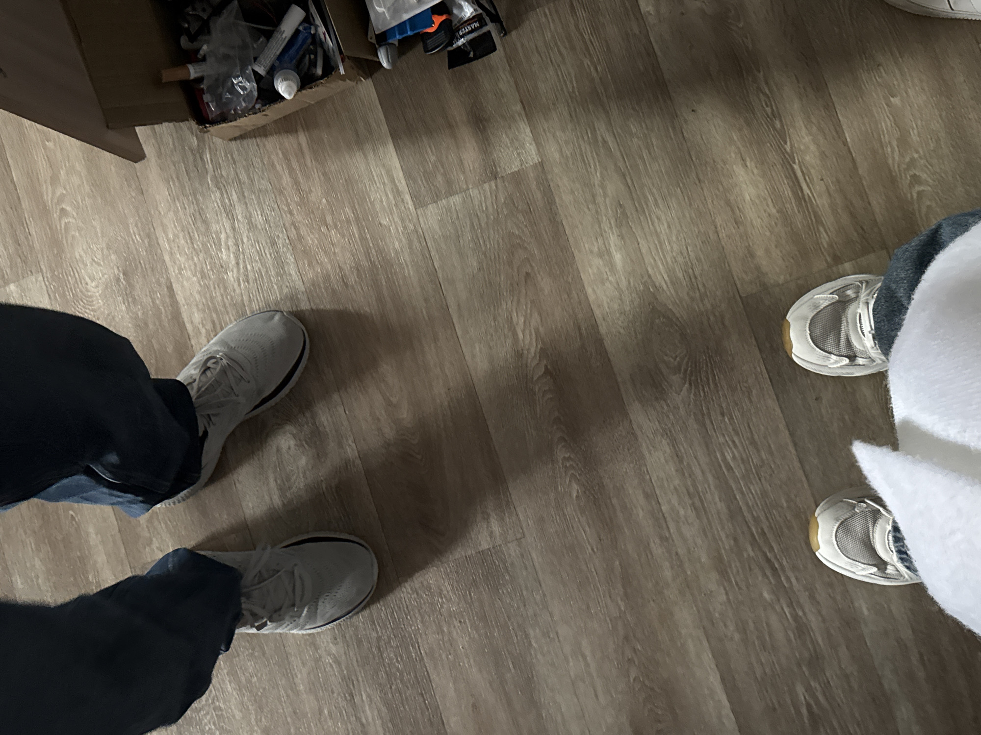 The image shows a section of a room with a light-colored wood-patterned floor. In the upper-left corner, there's a cardboard box containing various items. Two pairs of feet wearing sneakers are visible. One pair, located on the left side of the frame, is clad in grey sneakers and dark blue jeans. The other pair, situated in the upper-right, features white sneakers and is partially obscured by a white fluffy object. The scene appears to be indoors. The image doesn't provide information to determine its location within the city of Perm, Russia.