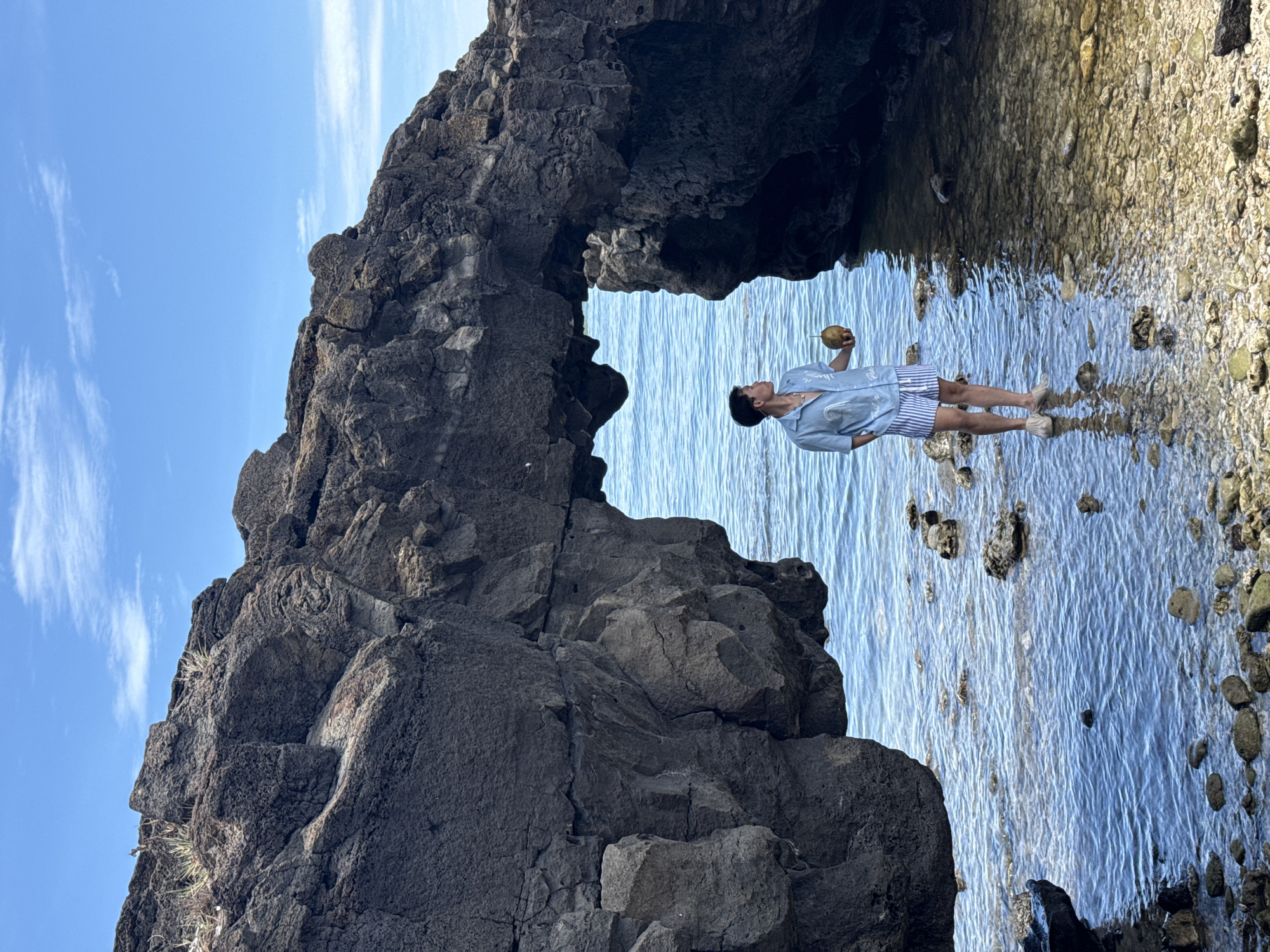 A person stands in shallow ocean water near a rocky arch formation. The person, facing away from the viewer and to the right, is holding a coconut. They are wearing a light blue short-sleeved collared shirt with a white graphic design and blue and white striped shorts, and light-colored shoes. The water is clear and shallow, with rocks and pebbles visible on the seafloor. The rocky arch, dark gray in color and irregular in shape, frames the ocean view. The sky above is blue with scattered white clouds. The scene appears to be on Lý Sơn Island, Quảng Ngãi Province, Vietnam.