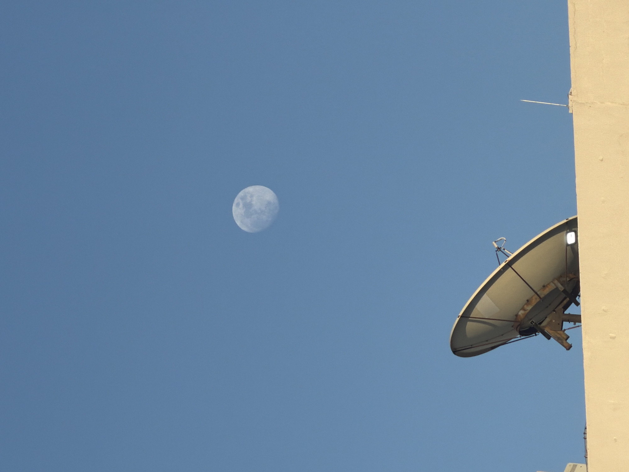 The image shows a daytime sky in Germasogeia Municipality, Cyprus. A full moon is visible in the upper-left quadrant. A satellite dish is mounted on the side of a building on the right side of the image. A thin lightning rod extends upwards from the building's edge. The sky is clear and blue.