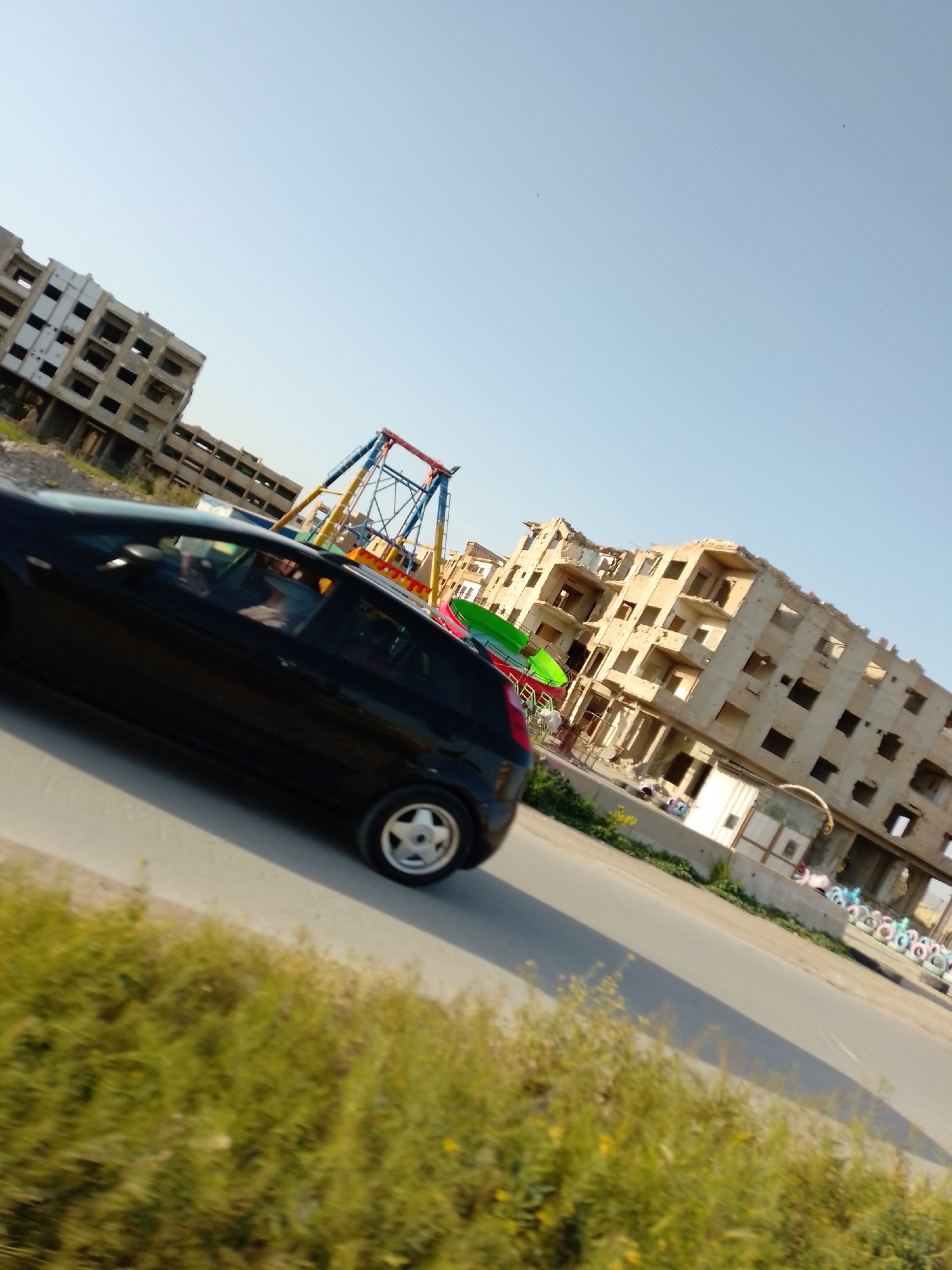 The image captures an outdoor scene featuring a black hatchback car, possibly a Fiat Punto, traveling on a paved road. At least two individuals are visible inside the car; one appears to be in the driver's seat and another in the front passenger seat, though their features are obscured.
To the right of the road, two large, multi-story buildings appear to be in various stages of construction or disrepair, featuring many empty window and door openings. Between the car and these structures, a prominent and colorful swing ride, characterized by blue, red, and yellow components, is visible, along with green and red plastic or fiberglass elements that suggest other amusement rides. Further in the background, near a low wall, a small white building with a dark roof stands, and a row of colorful painted tires lines the ground, possibly serving as a decorative barrier.
The foreground is occupied by a strip of green, overgrown grass and weeds bordering the road. The sky is clear and light blue, indicating bright, sunny daytime conditions. A long shadow cast by the car on the road suggests it is either morning or late afternoon. The scene combines elements of urban development or decay with a seemingly disused recreational area. No discernible text is visible anywhere in the image.