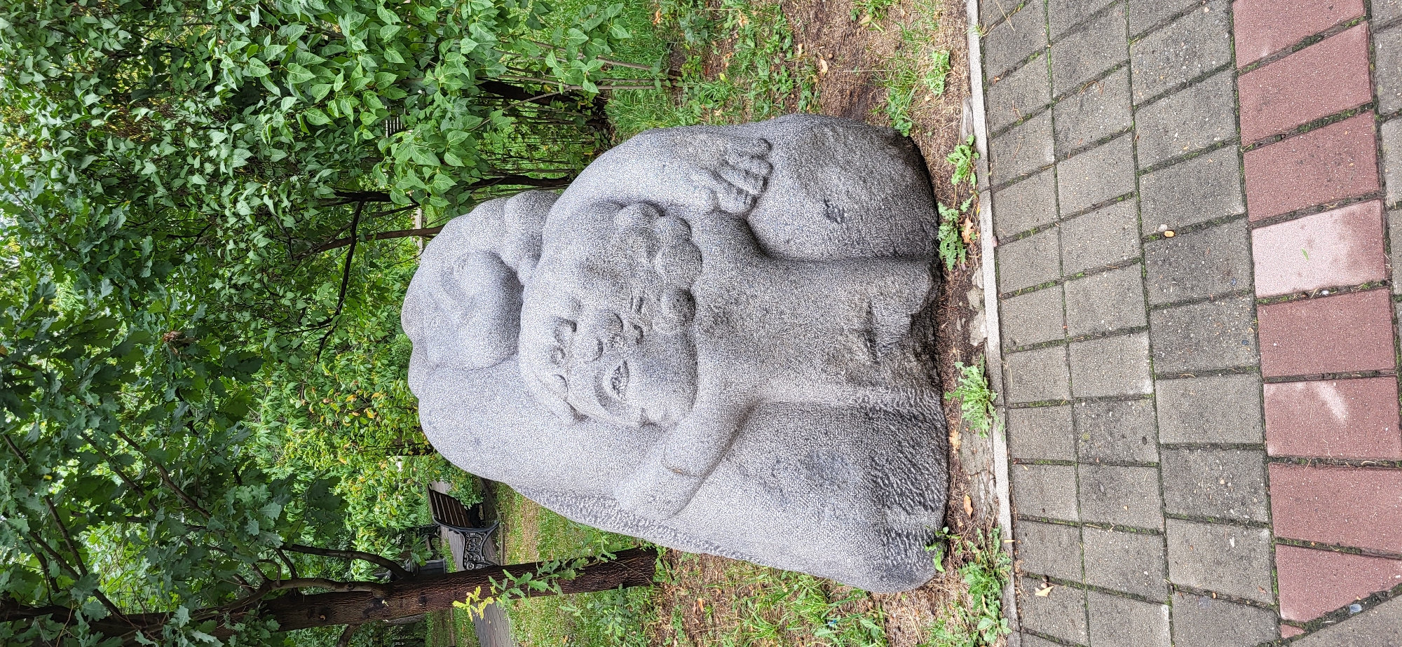 A grey granite sculpture of a mother and child is situated in a park in Oskemen, Kazakhstan. The sculpture depicts a large, rounded form, presumably the mother, cradling a smaller figure, the child. The child's face is visible, with eyes, a nose, and a mouth etched into the stone, and their arms are wrapped around the larger form. The sculpture rests on a patch of grass and dirt, bordered by a paved walkway made of grey and reddish-brown interlocking bricks. In the background, lush green trees and foliage create a natural setting, with a park bench visible further back among the trees. The overall scene suggests a public space dedicated to art and recreation.