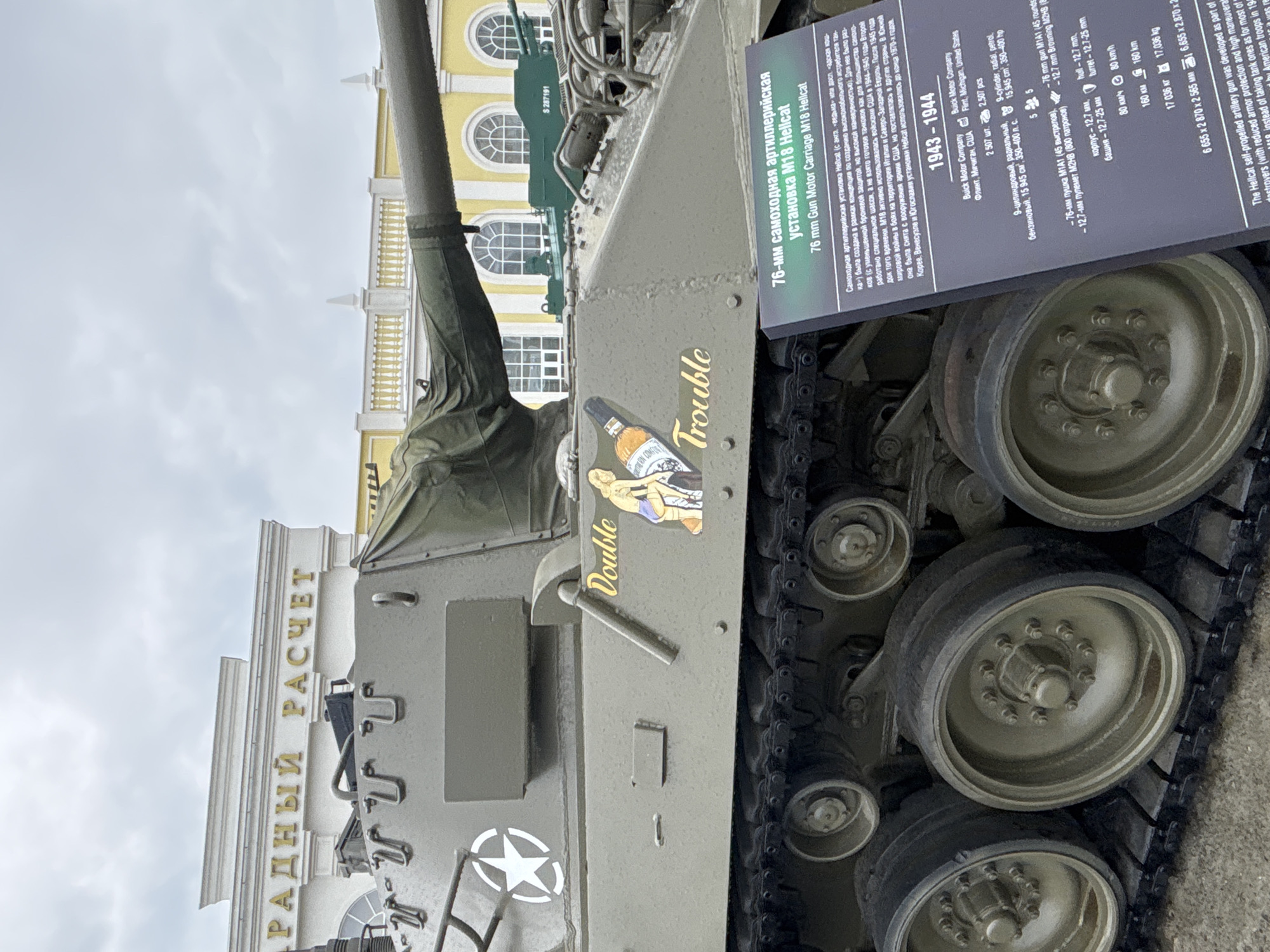 A close-up, slightly low-angle shot captures a U.S. M18 Hellcat self-propelled gun, painted olive drab, displayed outdoors. The tank's tracks, road wheels, and the lower portion of its hull are prominent in the foreground. A distinctive nose art decal featuring a blonde woman and a bottle is visible on the side of the hull, with the words "Double Trouble" written above and below it. The vehicle's long barrel gun extends across the frame. In the background, a portion of a classical-style building with arched windows and decorative molding is visible, along with a gray, overcast sky. A sign with both Russian and English text, detailing the M18 Hellcat's specifications and history, is placed in front of the tank. The sign indicates the tank's origin as the Buick Motor Company and its production during 1943-1944. The location is the Museum of Military Technology "Battle Glory of the Urals" in Verkhnyaya Pyshma, Russia.