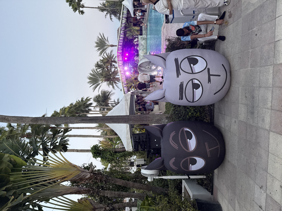 An outdoor evening event is depicted at the Fontainebleau Miami Beach resort in Miami Beach, United States of America. The scene is set at dusk, with the sky transitioning from a light blue to a pale purple hue.
In the immediate foreground, a light-colored paved stone walkway displays two large, approximately meter-tall, inflatable figures resembling stylized animal heads. The figure on the left is dark grey/black, and the one on the right is light grey, both featuring large eyes with a winking/smirking expression and a small smile. To the right of the light grey inflatable, a male individual with dark hair, wearing a patterned light-colored polo shirt, light pants, and dark shoes, sits on a low white structure. He is looking downwards, engaged in an action that appears to be cleaning an object with a small, light-colored brush. A second individual is partially visible standing on the far right.
Behind the dark grey inflatable, a white industrial stand-fan is mounted on a pole, and a stack of black sound equipment/speakers is visible amidst green foliage. The midground features a large swimming pool, reflecting purple and white lights originating from a decorated stage structure positioned at its far end. Figures, likely performers, are visible on the brightly illuminated stage, which is adorned with tropical greenery. Multiple white canopy tents are erected around the pool, housing guests who are seated or standing. Another white industrial fan is visible near the pool's edge.
The background is characterized by abundant tropical vegetation, including numerous tall palm trees of various species and several distinctive large Traveler's Palms with fan-like leaves on the left. Portions of multi-story white resort buildings are discernible through the dense foliage on the left side of the image.