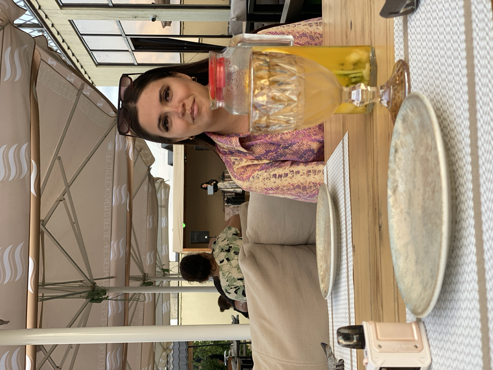 An outdoor dining scene in Derbent, Russia, features a woman seated at a table under a large, beige umbrella. The woman, with dark hair and wearing a patterned shirt, is looking towards the camera. In front of her is a table with two plates, silverware, and a glass pitcher filled with a yellow beverage and fruit slices. A vape device is also visible on the table. In the background, other patrons are seated at tables, and a stage with a performer can be seen. The setting appears to be a restaurant or cafe with an urban backdrop.