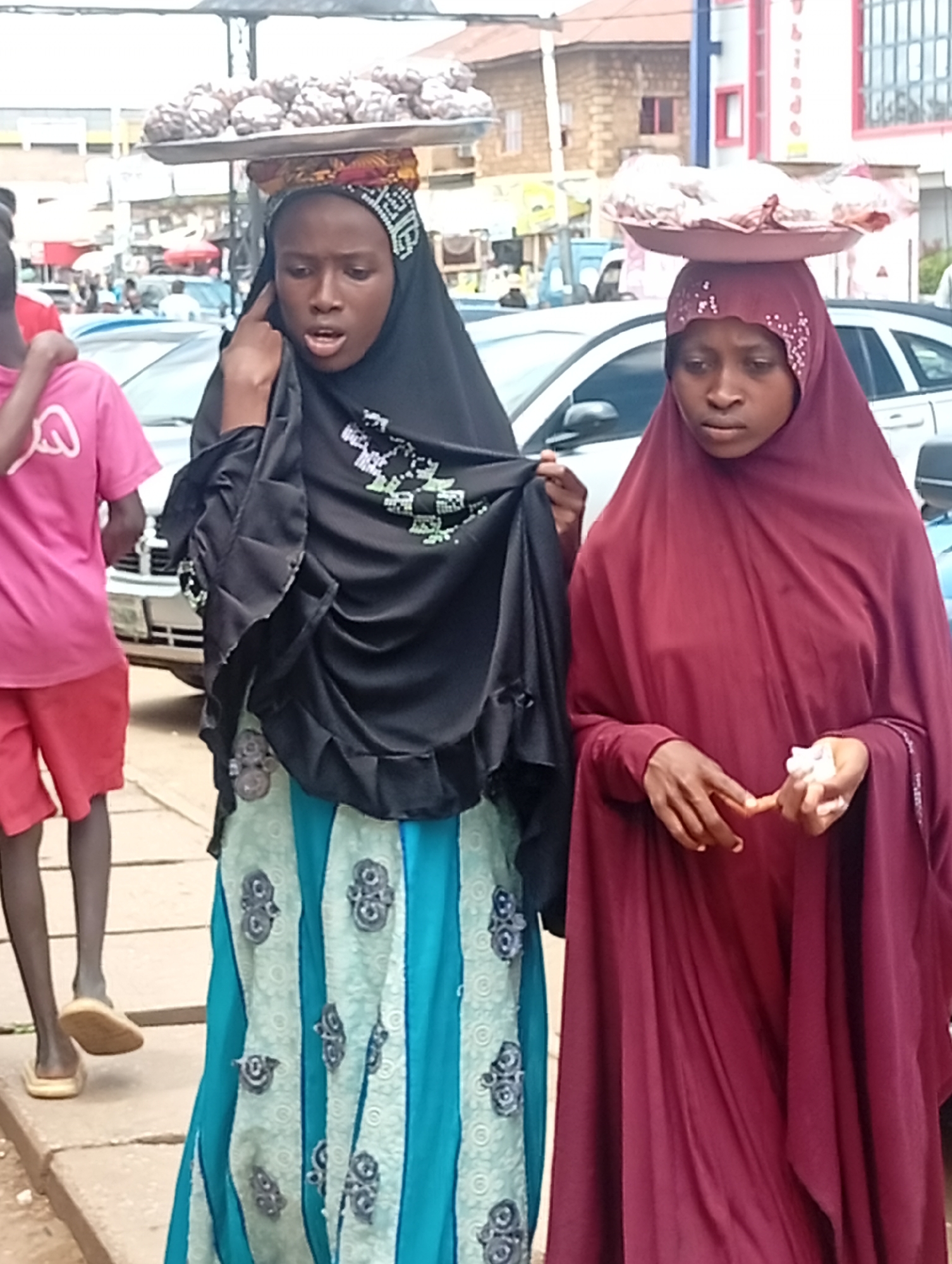 The image depicts two young women walking in a marketplace in Jos, Nigeria. The woman on the left wears a black hijab and a blue and white patterned skirt, carrying a tray of what appear to be baked goods on her head. Her mouth is open as if speaking or calling out. The woman on the right wears a maroon hijab and a matching dress, also carrying a tray of goods on her head, some of which are wrapped in plastic. She is holding something white in her right hand. In the background, vehicles are visible, suggesting a street or road adjacent to the market. The overall environment is an outdoor urban setting, likely bustling with activity.