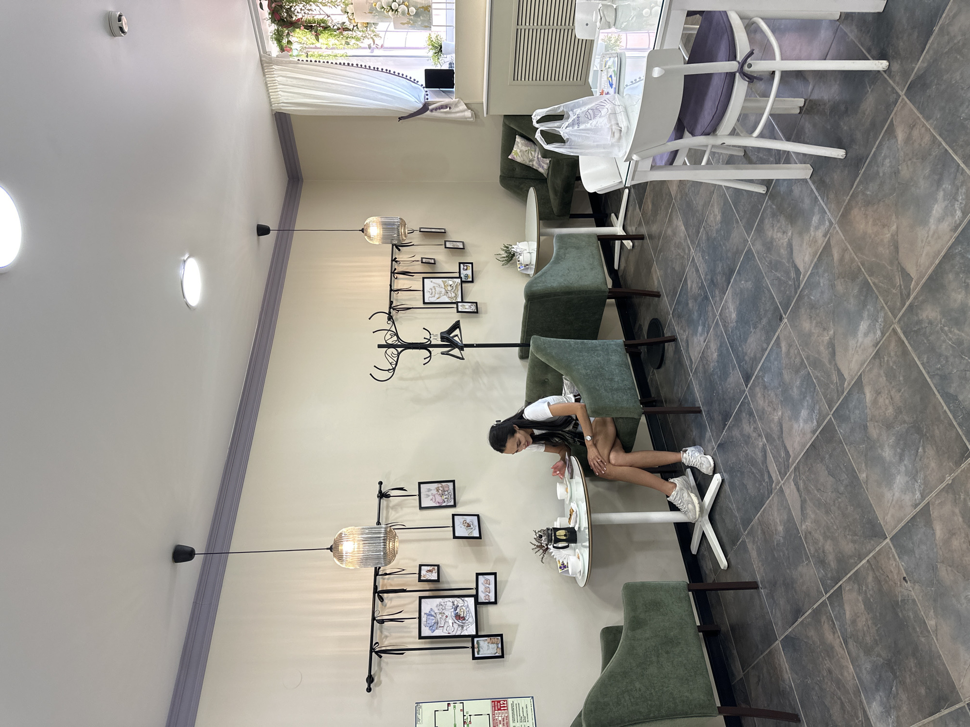 In a brightly lit cafe with a cream-colored ceiling and walls, a young woman with long dark hair is seated at a small round table. She is wearing a white t-shirt, black shorts, and white sneakers. Her gaze is directed downwards towards the table, where a few items are present, including what appears to be a cup and saucer. The table has a white top and a white pedestal base.
The cafe has a modern yet cozy ambiance. The floor is covered with large, square, dark gray tiles with a subtle marble pattern. Several plush green armchairs are arranged around the tables, creating intimate seating areas. A large window with sheer white curtains is visible in the background, suggesting it's daytime.
On the wall to the left of the woman, a decorative metal structure with various hooks holds framed pictures and what appear to be decorative ribbons. A large, ribbed glass pendant light hangs from the ceiling, illuminating the area. Further down the wall, a coat rack with several hangers stands empty. In the background, a white plastic bag is placed on a green armchair.
Visible text includes "112" on a red and white sign in the bottom right corner of the image. The overall scene conveys a sense of quiet and relaxation, as if the woman is enjoying a moment of solitude in the cafe. The lighting suggests it is daytime.