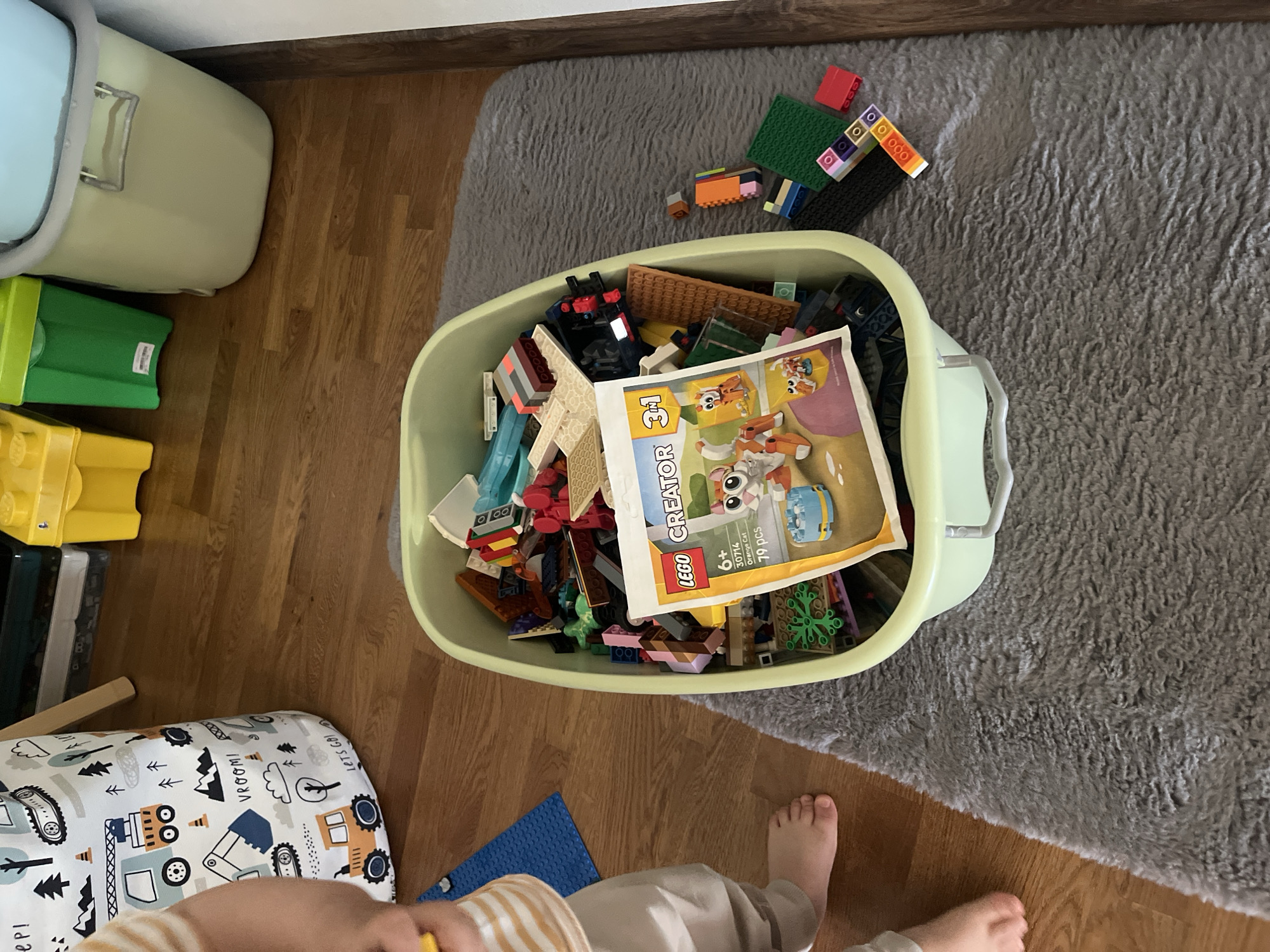 The image depicts a child's legs and feet on a wooden floor next to a fuzzy grey rug. On the rug is a light green plastic bin overflowing with Lego bricks, a Lego Creator instruction booklet, and other building toys. Scattered on the rug outside the bin are additional Lego pieces. In the background, a large white fabric storage bag with a vehicle pattern is visible, as well as stacks of yellow and green plastic containers. The scene appears to be indoors, likely a playroom or living area. There are no explicit indicators of the city of Onokivtsi, Ukraine, in the image.