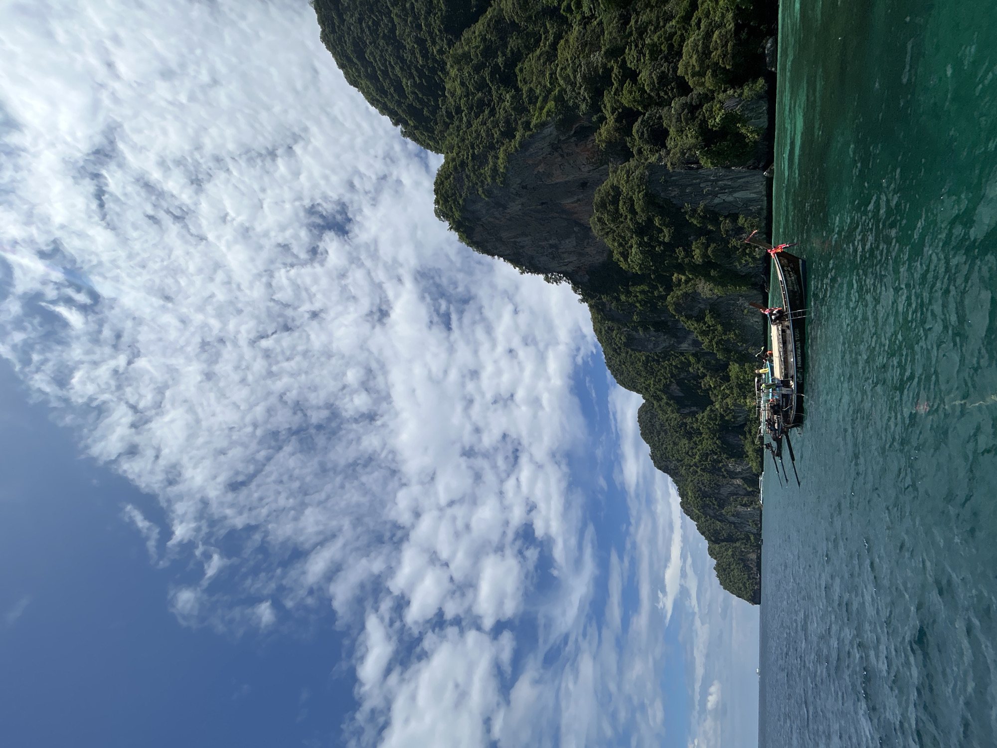 This is a scenic outdoor photograph taken from a boat in the waters off the coast of Ao Nang, Thailand. The image captures a majestic limestone cliff covered in lush green vegetation, rising from the turquoise waters. The sky above is a vibrant blue, dotted with scattered white clouds. Several traditional Thai longtail boats are visible moored near the shore, some with people on board. The water is clear and calm, with the seabed visible in some areas. The scene evokes a sense of tropical beauty and tranquility.