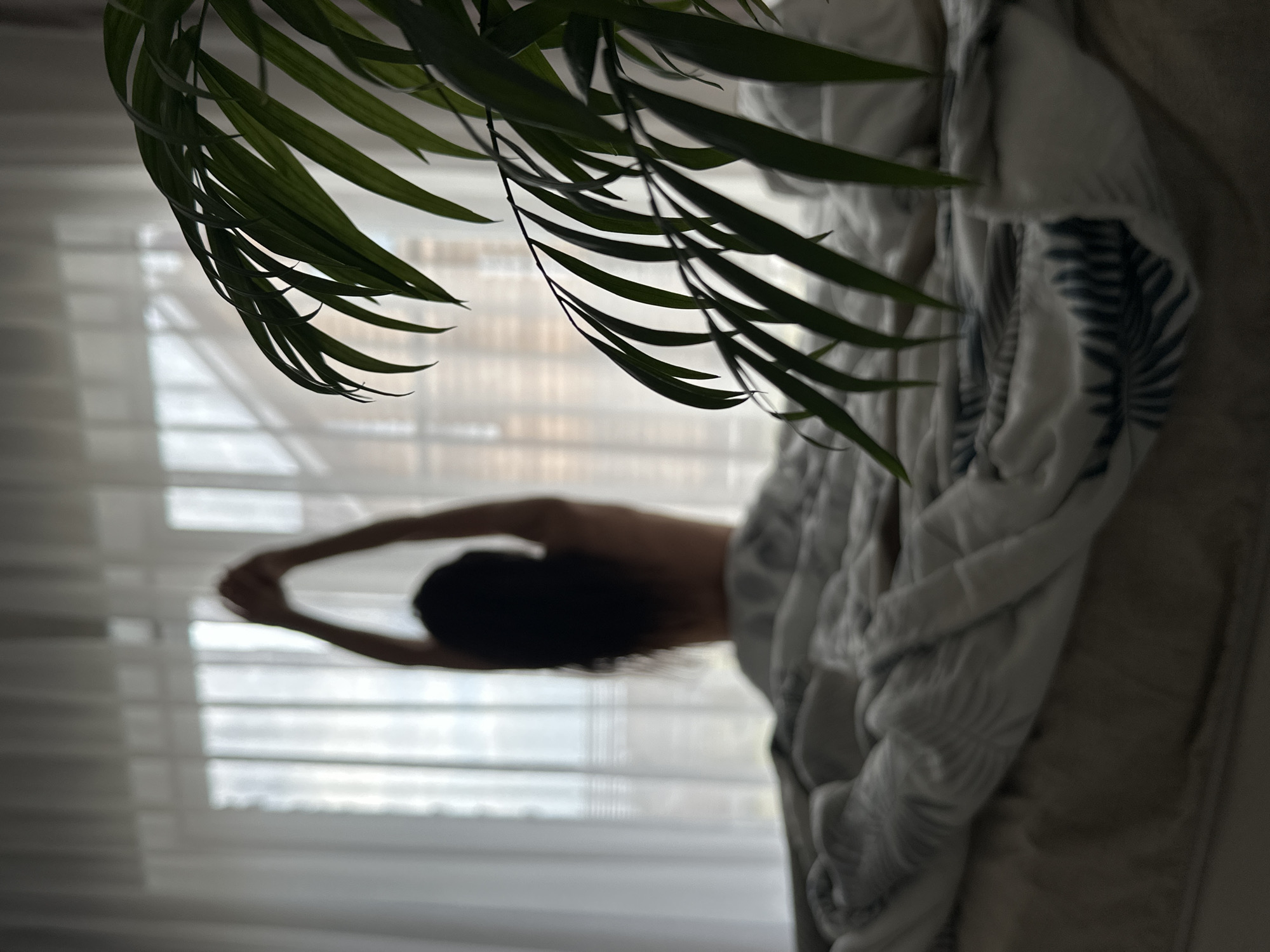 The image depicts a person in a room with a plant and a blanket. The person, whose features are blurred, appears to be stretching their arms overhead. They are in front of a window with sheer curtains. A leafy green plant hangs in the foreground. A patterned blanket is draped on what seems to be a piece of furniture. The location is suggested to be Chaikovsky, Russia, however no element present in the image validates this.
