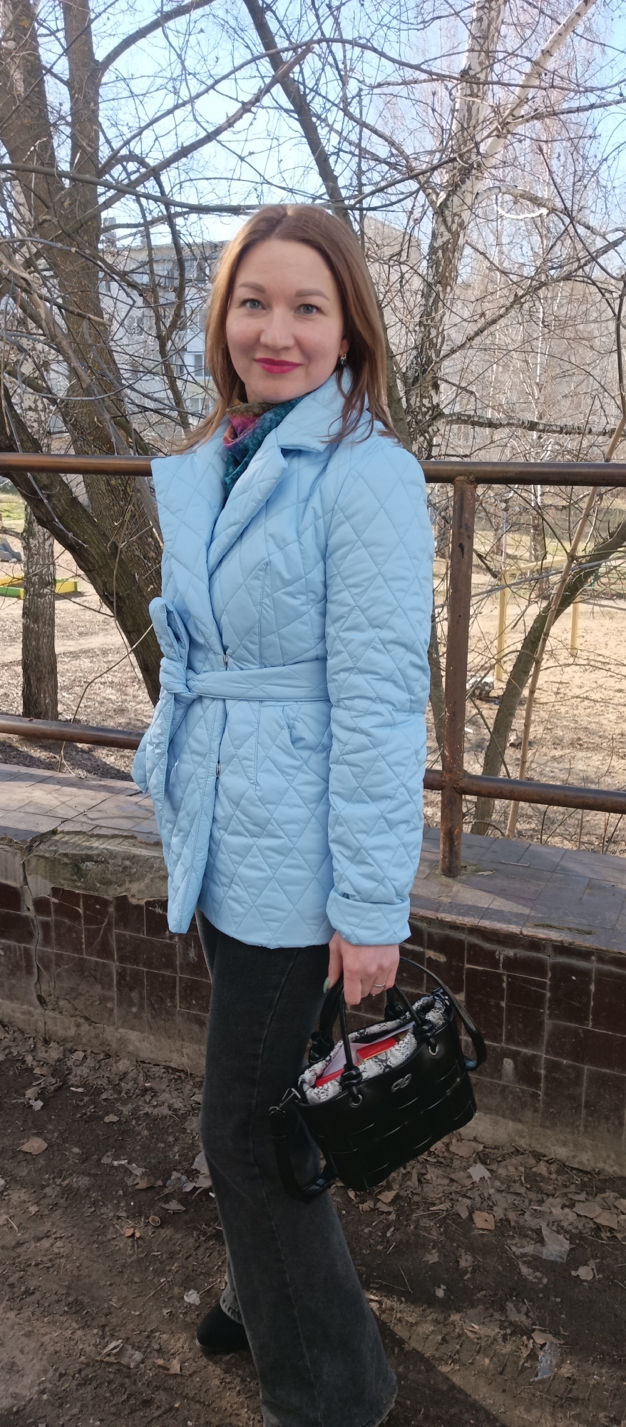 A woman is standing outdoors next to a low brick wall with a metal railing behind her. She is wearing a light blue quilted coat with a tie waist, dark jeans, and holding a black handbag. The background features trees without leaves, suggesting it is autumn or winter. The location is Unknown, Unknown.