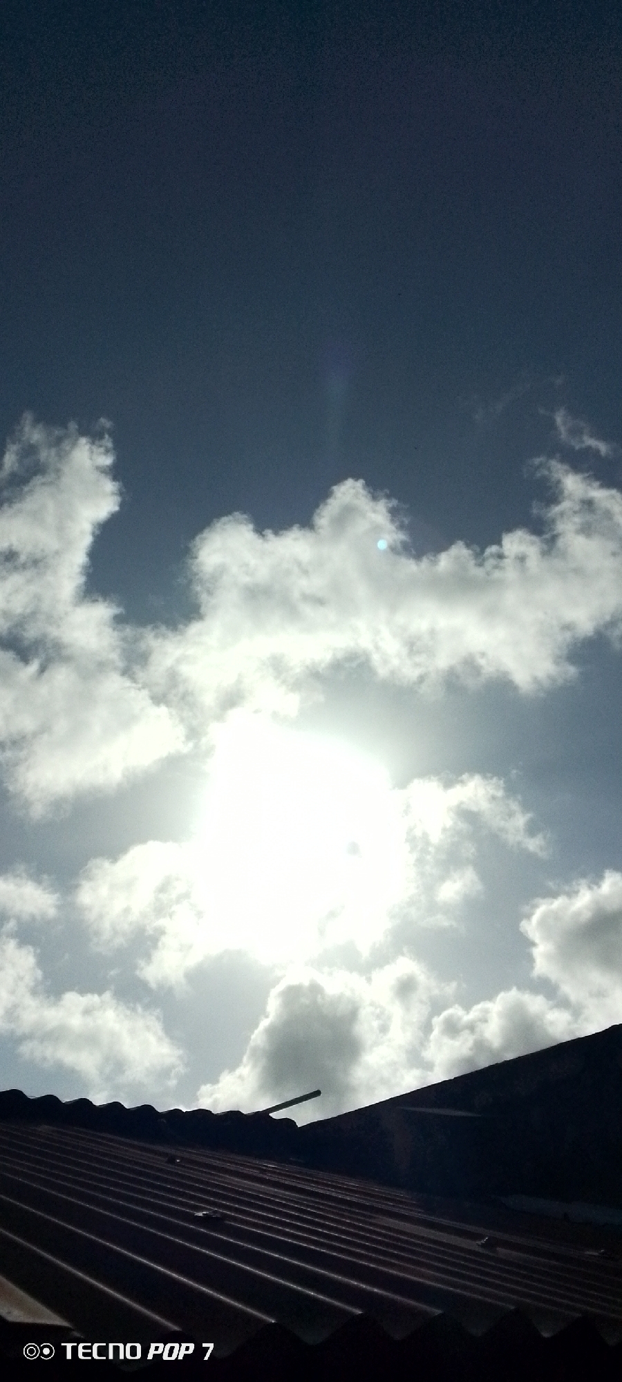 The image shows a partly cloudy sky above a corrugated iron roof. The sun is shining brightly behind the clouds, creating a hazy glow. The sky is blue, though partially obscured by the clouds. A portion of the roof is visible in the lower part of the image. The image was taken with a TECNO POP 7 mobile phone. The location, Altagracia, Venezuela, is based on the prompt, and is not evident from the image itself. No people, subjects, actions, or interactions are visible.
