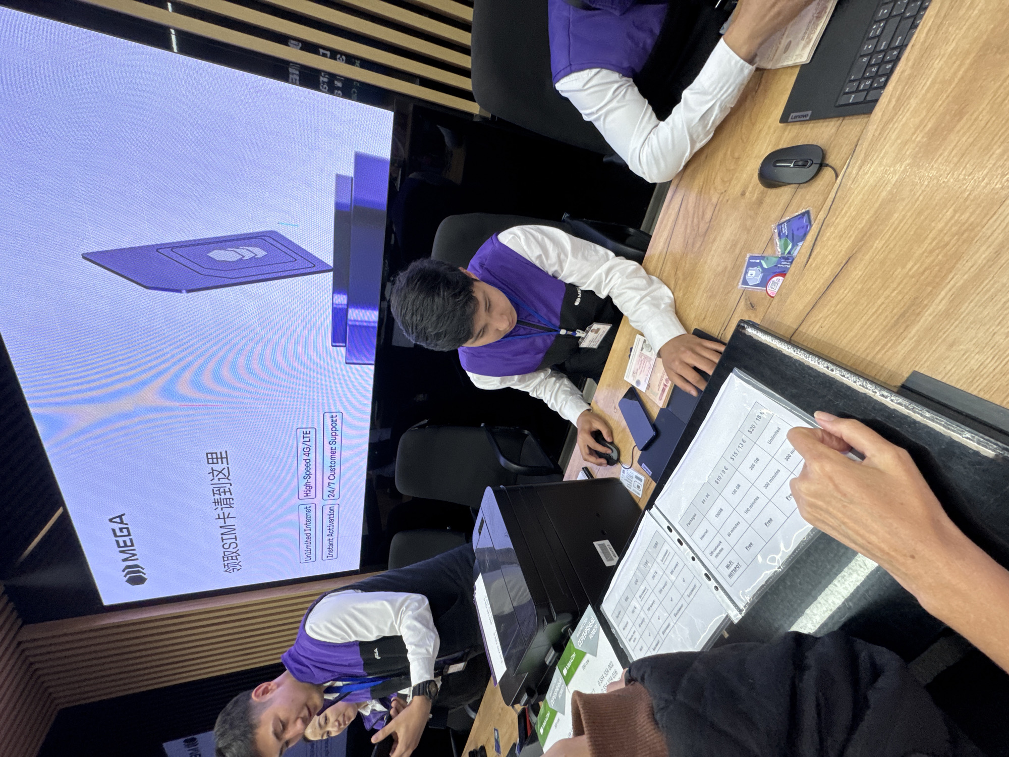 The scene appears to be inside a MegaCom mobile service center in Bishkek, Kyrgyzstan, depicting interactions between staff and customers.
There are at least two employees wearing white shirts with purple vests. One employee is assisting a customer, using a computer and possibly a service catalog with tariff information. A second employee is helping another customer. A large screen in the background displays the MegaCom logo and advertising for SIM cards, highlighting features like "Unlimited Internet," "High-Speed 4G/LTE," "Instant Activation," and "24/7 Customer Support." The text on the screen is also in Chinese. The table is wooden. A black Lenovo keyboard is visible. A notebook of service packages is visible with pricing in both dollars and Euros.