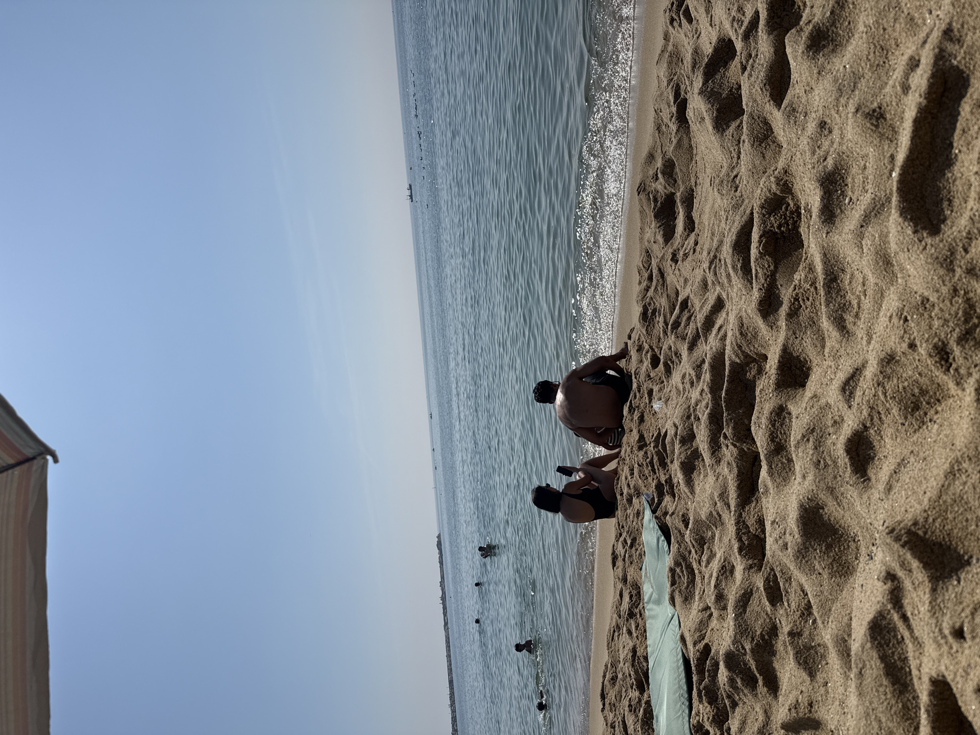 This is an eye-level, medium shot of a beach scene on a clear day, likely during the summer. In the foreground, a sandy beach is covered in undulating dunes, with a green mat placed on the left side. Two individuals, a man and a woman, are seated on the sand near the water's edge, facing away from the camera. The woman, wearing a black swimsuit, holds a smartphone. The man, shirtless and wearing dark swim trunks, is also looking out at the water. The sea is calm with gentle waves lapping at the shore, creating a shimmering effect. In the middle ground, several people are swimming and playing in the water. Further in the distance, a breakwater extends into the sea, with a few boats visible on the horizon. The sky is a clear, pale blue, with the top edge of what appears to be a striped beach umbrella visible at the very top of the frame. The overall impression is one of a relaxed beach day. Given the visual cues such as the architecture of the breakwater and the general atmosphere of a Mediterranean beach, this scene is likely located on the coast of Barcelona, Spain.