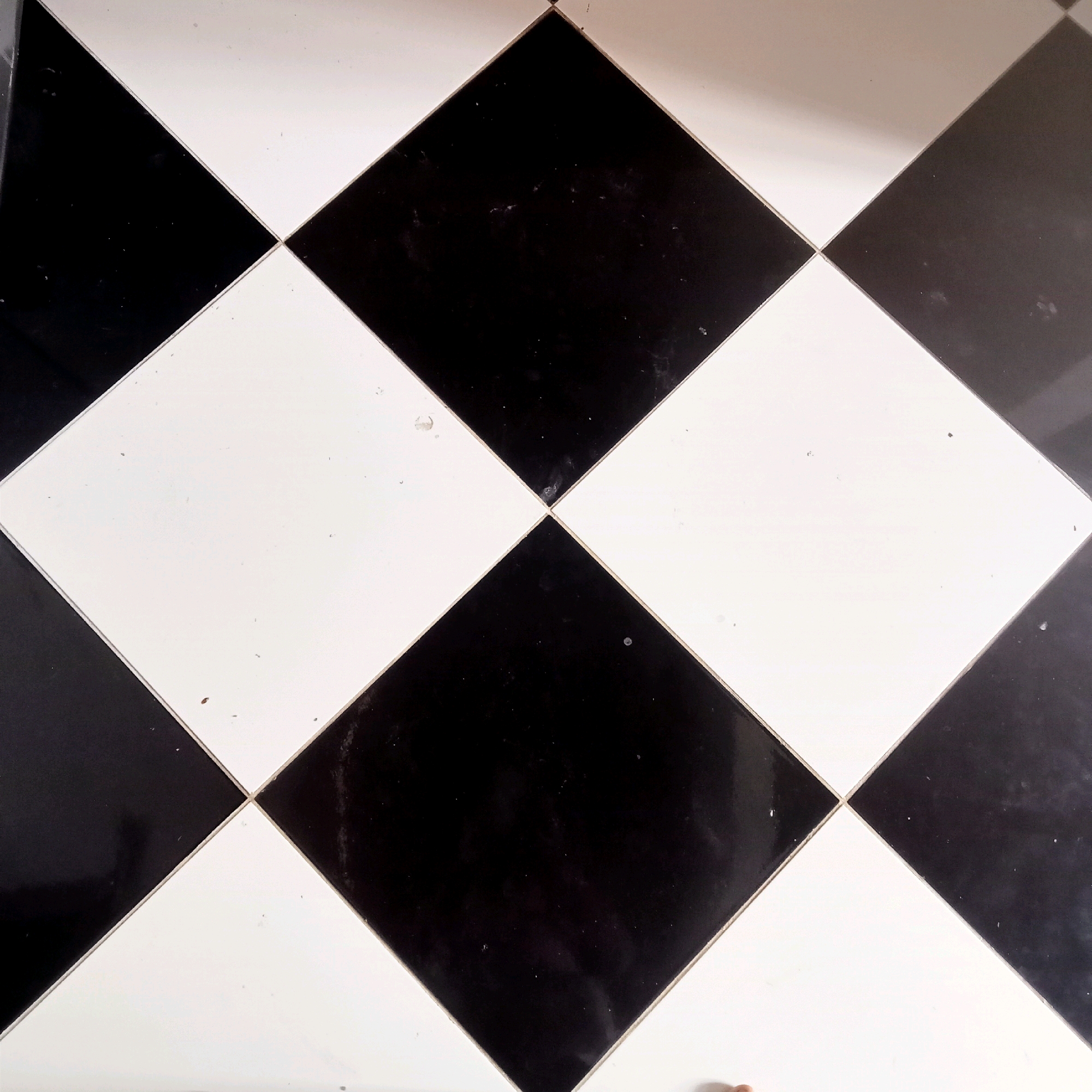 The image shows a close-up, top-down view of a black and white checkered floor. The tiles are laid in a diamond pattern, with the points of the squares meeting in the center. The floor appears to be made of a glossy material, possibly ceramic or polished stone, as evidenced by the reflections on the surface. There are a few small specks of dust or debris visible on both the black and white tiles, suggesting the floor may not be perfectly clean. The lighting in the image is somewhat uneven, creating subtle highlights and shadows on the glossy surface. There is no visible context that would indicate the location in Jaten, Indonesia.