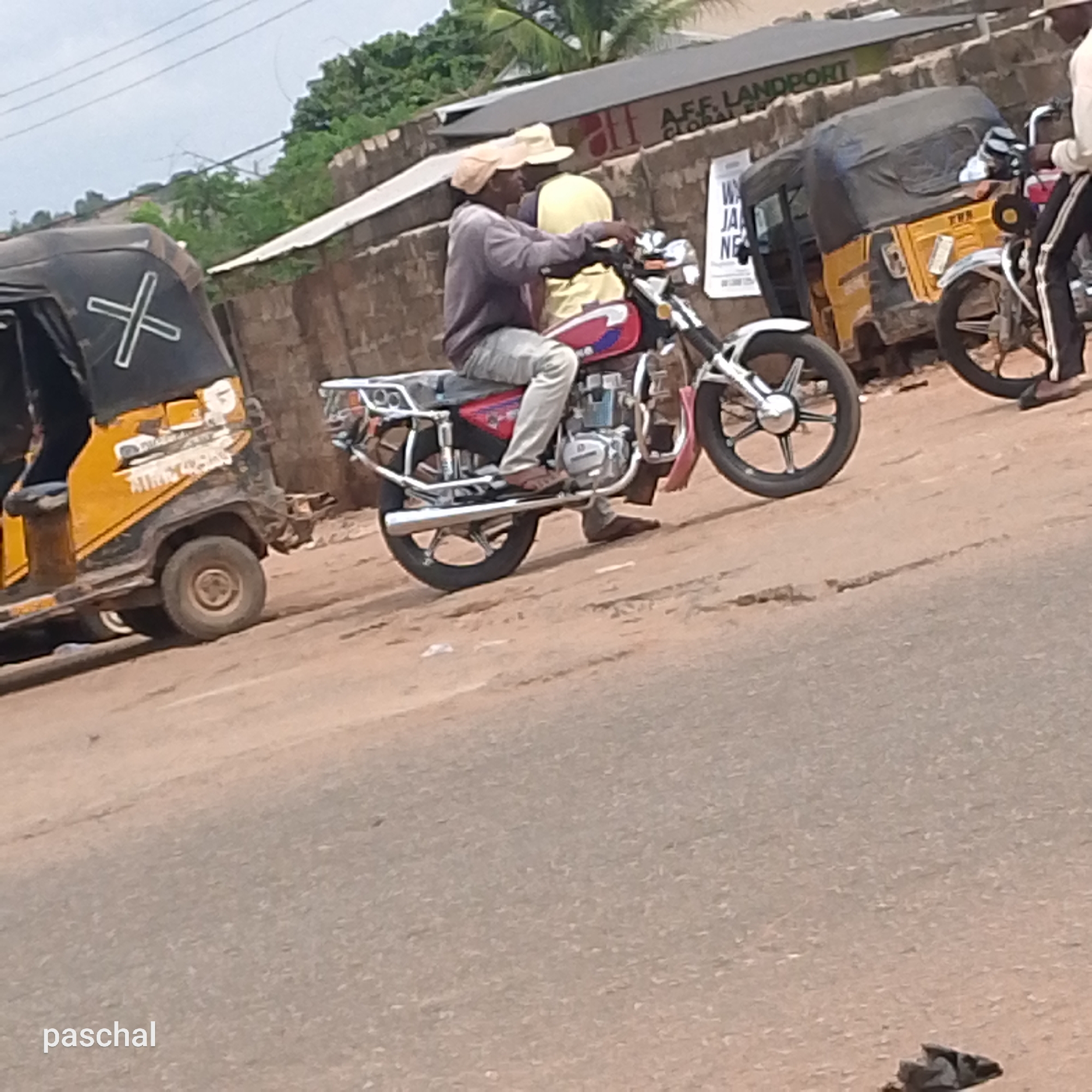 A man wearing a hat is riding a motorcycle with a passenger in Nsugpe, Nigeria. Nearby are auto rickshaws, a building with a sign, and a dirt road.
