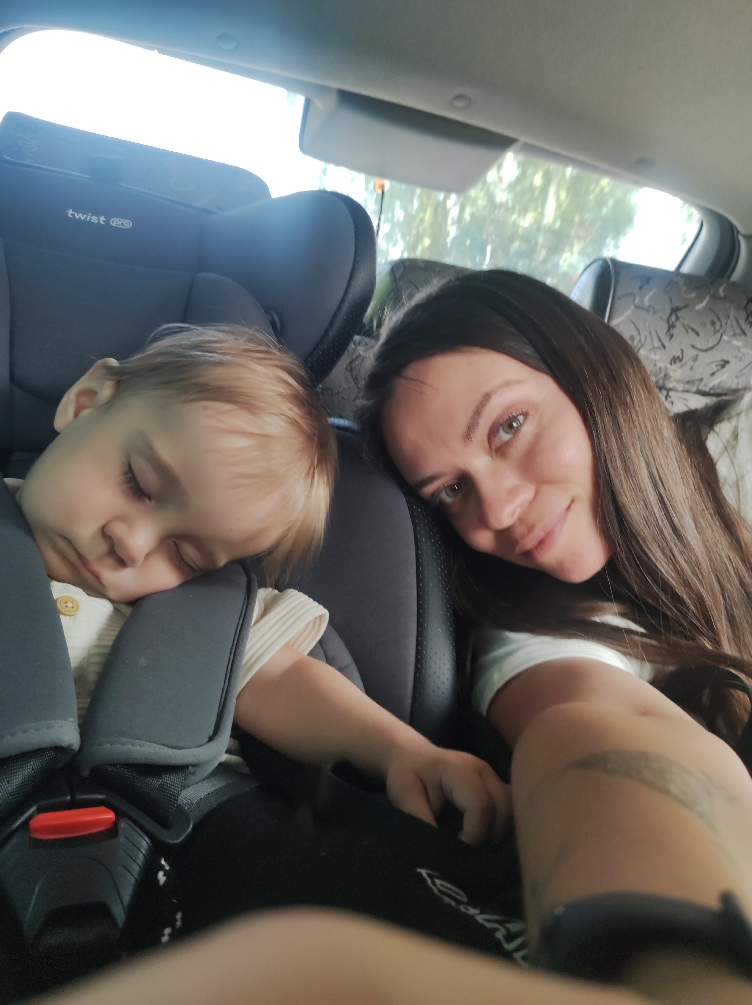 The image depicts a woman and a sleeping child in a car, likely in the back seat. The child, with blond hair, is strapped into a gray car seat with a "twist" logo visible on the headrest. The woman, with long brown hair and fair skin, is leaning in close to the child, appearing to take a selfie. The car's interior includes patterned headrests and a view out the window suggesting trees and daylight. There's no definitive evidence that indicates this image was taken in Chaikovsky, Russia.