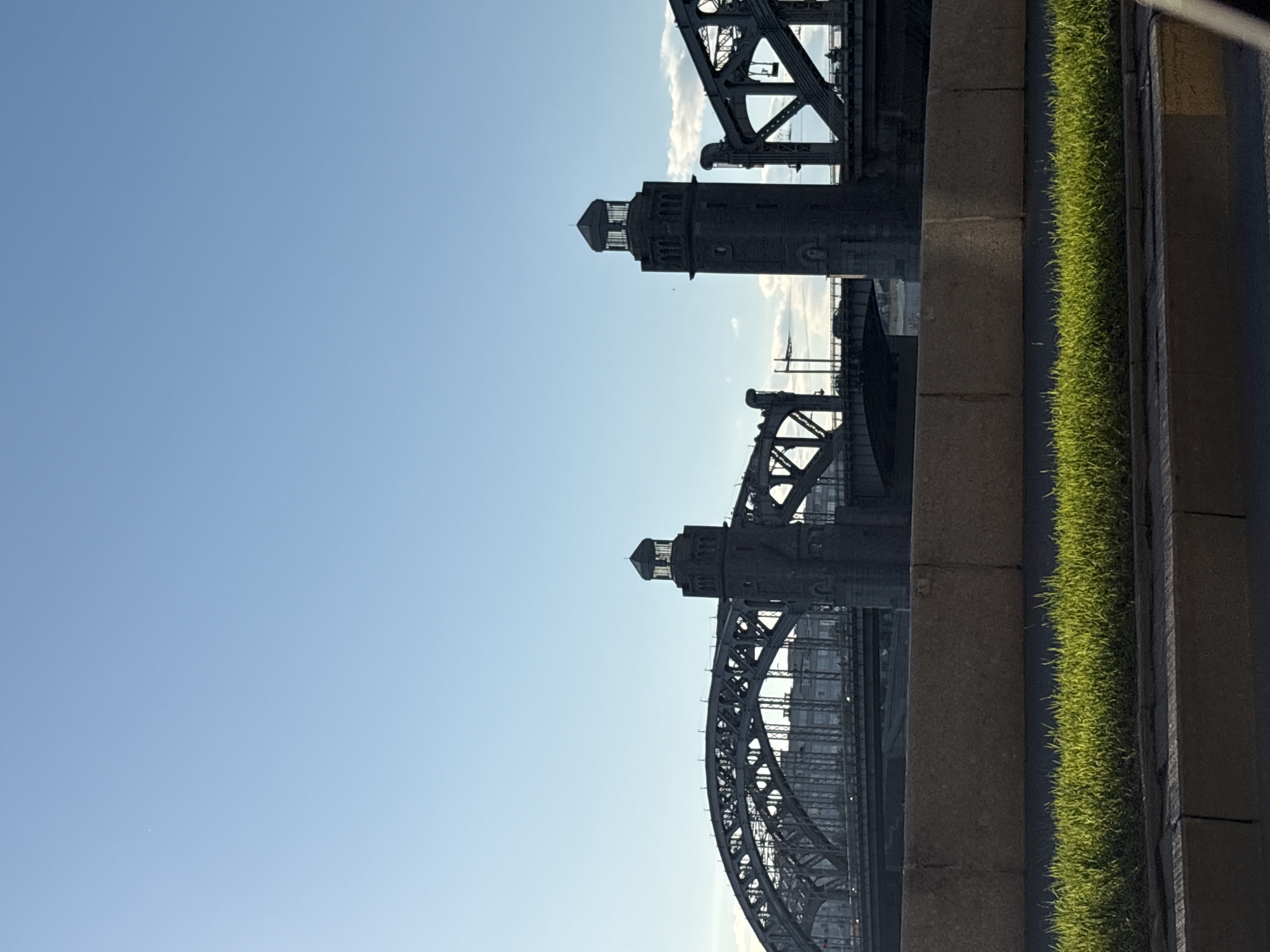 The image depicts a view of the Bolsheokhtinsky Bridge in Saint Petersburg, Russia, from a low-angle perspective. The sky is a clear, pale blue. The bridge's steel structure is visible, along with its ornate towers. A concrete retaining wall with a strip of green grass is visible on the right side of the frame.