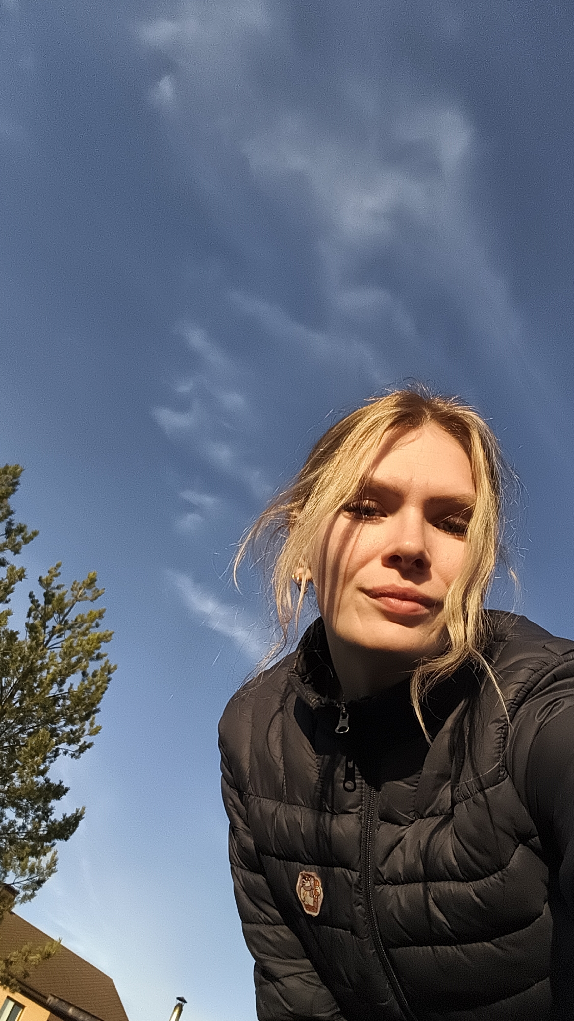 A low-angle outdoor selfie features a young woman in Русский Вожой, Russia, looking directly at the camera. She has light blonde hair, parted in the middle with strands falling across her forehead and framing her face. She is wearing a black, quilted puffer jacket, and on the right side of her chest, there is a small, unidentifiable badge or patch depicting a stylized character, possibly an animal. Her expression is calm, with a subtle, slight smile.
The background is dominated by a clear, bright blue sky with scattered, thin white clouds. The warm, directional lighting suggests the photo was taken during either the late afternoon or early morning, casting soft shadows on her face and highlighting her hair. To the left, a portion of a vibrant green coniferous tree is visible, and below it, the brown roof and cream-colored wall of a building are partially seen. The overall setting is sunny and pleasant, capturing an ordinary moment outdoors. No discernible text is visible on the badge or elsewhere in the image.