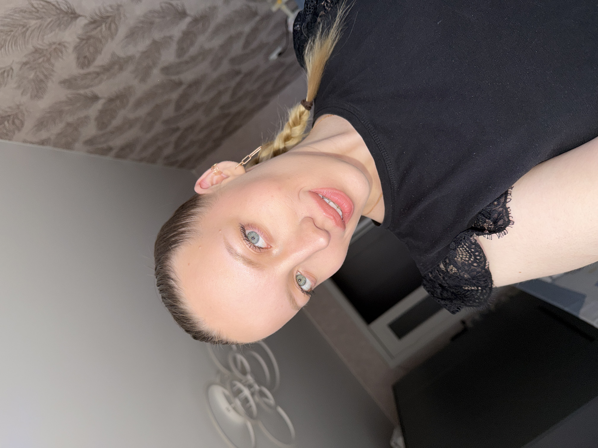An adult female is depicted in a close-up indoor selfie, assumed to be located in Reshetnikovo, Russia. She has fair skin, a subtle smile revealing upper teeth, and her gaze is directed at the camera. Her left eye appears light blue-green, and her right eye appears light blue, indicating heterochromia. Her hair, dark brown at the roots and lighter blonde through the lengths, is pulled back, with a portion braided into a single plait secured by a brown elastic. She wears a black short-sleeved top with lace detailing on the sleeves and a thin gold-toned necklace. The visible right ear features a small gold-toned ear cuff and a linked gold-toned drop earring. The background includes a white ceiling with a modern, multi-ringed light fixture in the upper left, a dark, possibly black, door frame or furniture piece partially visible on the left, and a wall covered in patterned wallpaper featuring a subtle feather or leaf motif on the right.