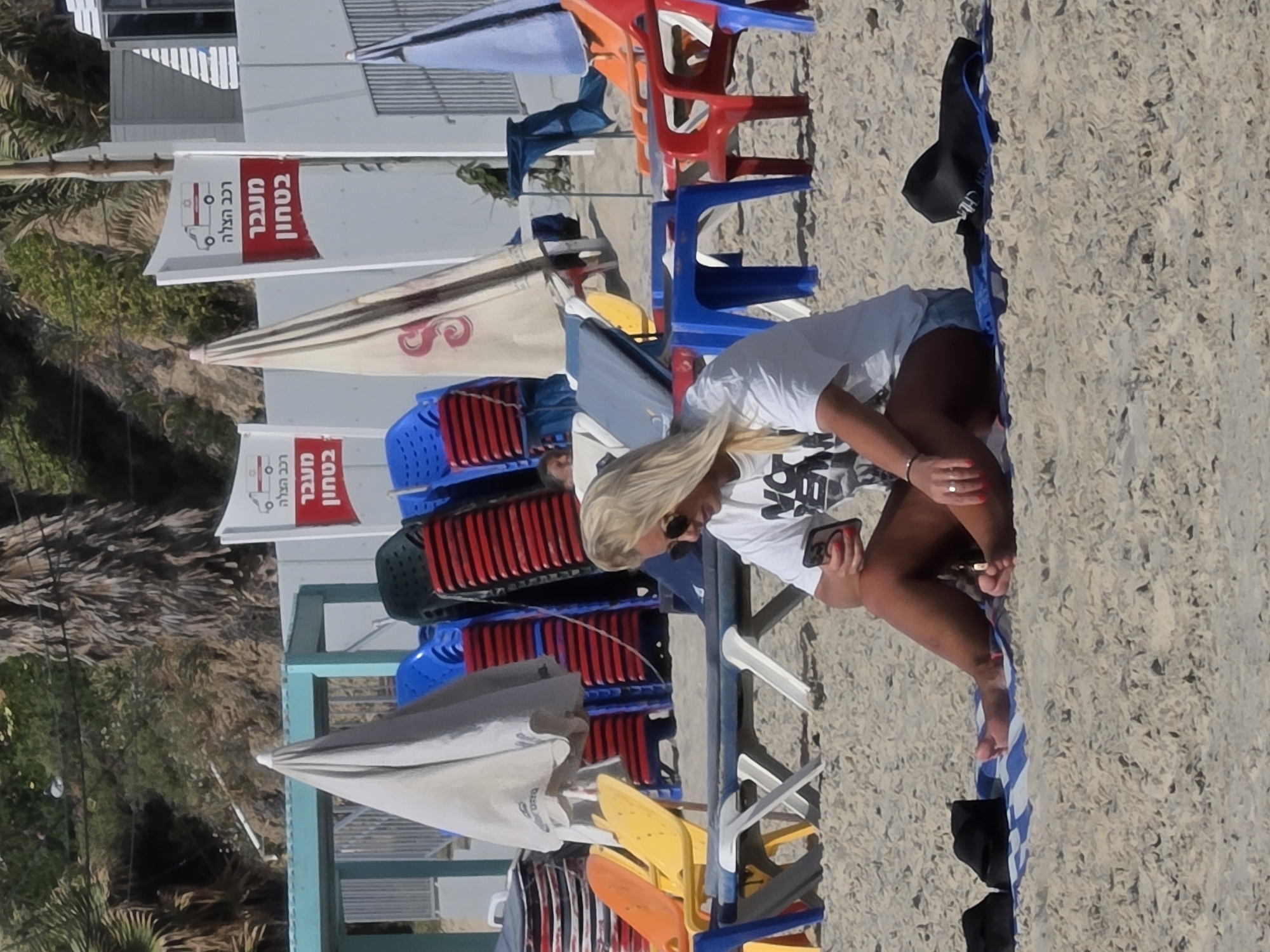 This image depicts a sunny beach scene, likely in Bat Yam, Israel, given the contextual information. The central subject is a woman with long, blonde hair, wearing a white t-shirt, denim shorts, and sunglasses. She is seated on a white plastic beach chair, engrossed in her mobile phone, which she holds in her left hand. Her fingers and toes are adorned with red nail polish, and a ring is visible on her left ring finger.
The beach itself is covered in light-colored sand. Scattered across the scene are numerous colorful plastic beach chairs and umbrellas, suggesting a public or commercial beach. The chairs come in various shades including red, blue, yellow, orange, white, grey, and black, with many stacked neatly. Several beach umbrellas are present, including a cream-colored one with red detailing and a grey one. To the right of the woman, a blue and white striped beach towel lies on the sand, accompanied by a black hat.
In the background, white buildings with grey horizontal shutters are visible, suggesting beach facilities or nearby structures. Lush green foliage, including several palm trees, provides a tropical backdrop. Two distinct red signs with white Hebrew text are prominently displayed on the buildings. The text on these signs reads "בטחון" (Security/Safety), "מעבר" (Passage/Crossing), and "רכב הצלה" (Rescue Vehicle), accompanied by an ambulance icon. This signage indicates a designated path or access point for emergency vehicles, likely near a lifeguard station. The overall atmosphere is one of relaxation and leisure on a bright, clear day.
