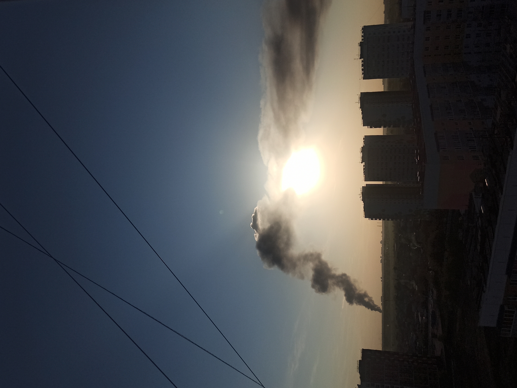 The image depicts a cityscape in what appears to be Кондратово, Russia. Silhouetted apartment buildings line the right side of the frame. In the distance, dark smoke rises from the ground, obscuring the sun and creating a haze in the sky. Electrical wires stretch across the lower left of the image. The sky transitions from dark blue on the left to a hazy orange near the horizon.