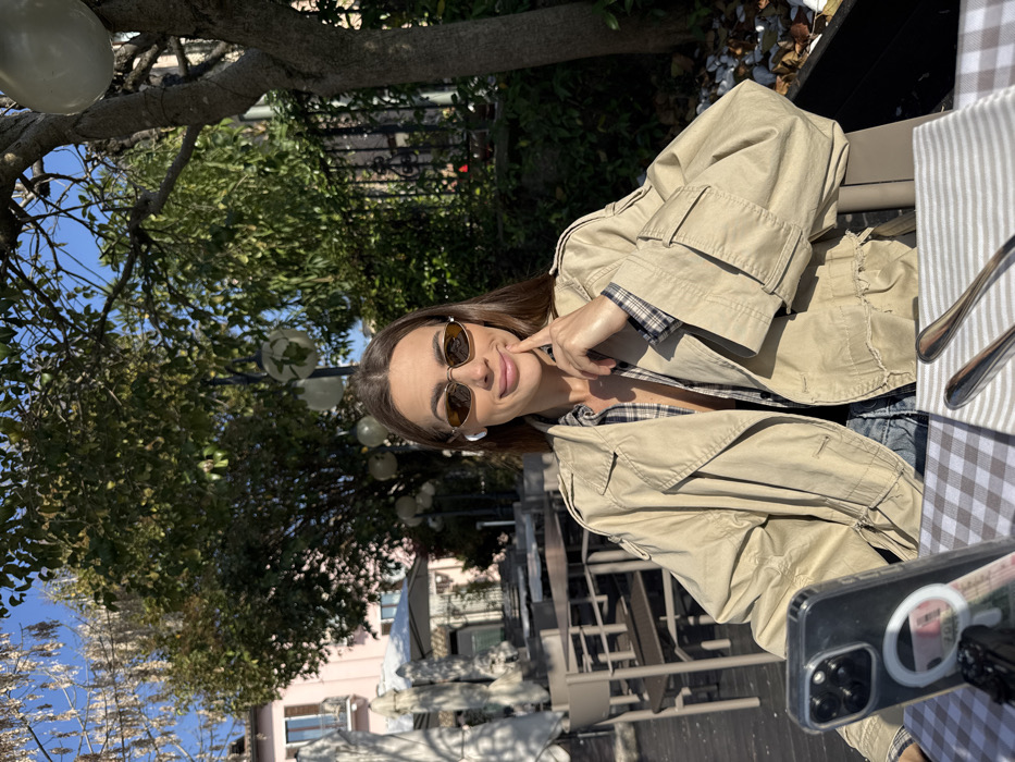 A female subject is depicted seated at an outdoor dining table within a cafe setting in Sirmione, Italy, under clear daylight conditions. She has long dark hair, wears oval sunglasses, and a white earbud in her left ear. Her attire consists of a light tan trench coat worn over a plaid button-up shirt. Her left hand is raised, with fingers resting against her chin. The table in front of her is covered with a brown and white checkered tablecloth. An upright smartphone, identified as an iPhone series with a MagSafe ring on its clear case, is positioned on the table's left side; a pink 20 Euro banknote is partially visible behind it. To the right, a folded white and grey striped napkin holds a fork and knife. The background features an outdoor seating area with multiple unoccupied grey tables and chairs, white patio umbrellas, and mature trees with green foliage. Several spherical white lights are suspended from the tree branches. In the upper left background, a light pink building with windows and balconies is visible against a clear blue sky.