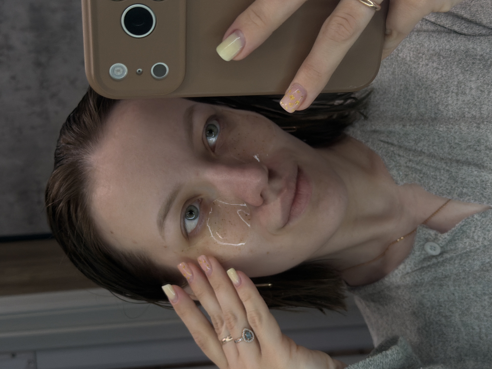 A woman is taking a selfie. She holds a brown smartphone in her right hand, partially obscuring her face. Her left hand is near her cheek. A clear, viscous substance is applied to her face in the area around her cheekbone and under her left eye. She has light skin, freckles, and short dark hair. She is wearing a gray sweater and a thin gold necklace. She has two rings on her left hand and her nails are painted with a yellow ombré effect on some fingers and a gold glitter design on others. The background appears to be a blurred wall with a gray tone. There is no indication of the location being Reshetnikovo, Russia.