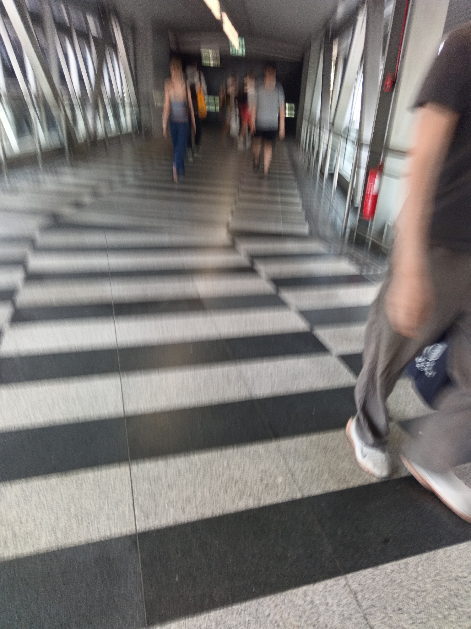 The image depicts an interior walkway or skybridge with a distinctive black and white striped floor pattern. The walkway appears to be enclosed by modern architecture featuring angled metal supports and glass panels. In the mid-ground, several individuals are visible, blurred due to motion. On the left, a woman in a gray tank top and blue pants walks away from the camera. To her right, a man in a gray t-shirt and black shorts is also walking away. Further back, other figures are less distinct. In the foreground, a person's legs and feet are prominently featured, with one foot wearing a white sneaker and the other foot out of frame. The overall impression is one of movement and transit within an urban environment. This scene is highly consistent with the architectural style and pedestrian overpasses commonly found in Kuala Lumpur, Malaysia, such as those connecting shopping malls or transport hubs.