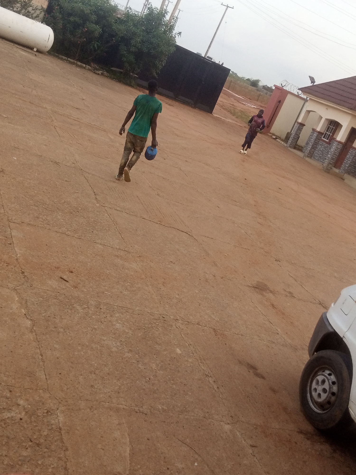 In an outdoor, urban environment, two young males are observed walking across a dusty, paved area. The individual in the foreground, wearing a green t-shirt and brown trousers, is walking away from the camera, carrying a blue spherical object in his right hand. To the right and further in the background, another male, dressed in a patterned shirt and dark trousers, walks towards the camera. A large white cylindrical tank is visible on the left side of the image, with a black gate and buildings in the background. The sky is overcast, and a white vehicle's wheel is partially visible in the lower right corner of the frame. The ground is a light brown, dusty surface with visible cracks.