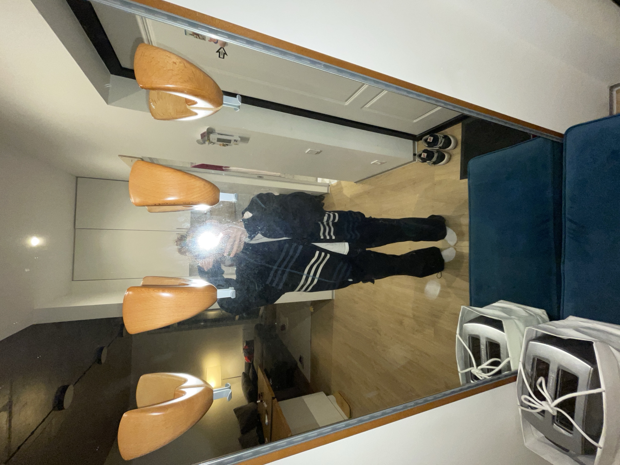 The image depicts a full-length mirror reflecting a person standing in what appears to be a hallway or entryway. The person, seen from the chest down, is wearing a dark jacket with white stripes and dark pants, with white socks visible. Their face is obscured by the reflection of a bright light source.
In the foreground, two white boxes with white string tied around them are placed on a dark teal bench. To the right of the bench, a pair of black and white sneakers are visible on the floor.
The mirror itself has a wooden frame with a metallic border. Mounted on the wall above the mirror are three wooden light fixtures with a curved design.
The reflection in the mirror shows a glimpse of a living space with furniture, including a sofa and a table, and a lamp. The floor is made of light-colored wood. The environment appears to be indoors. There is no visible information in the image to indicate the location is in the city of Khimki, Russia.