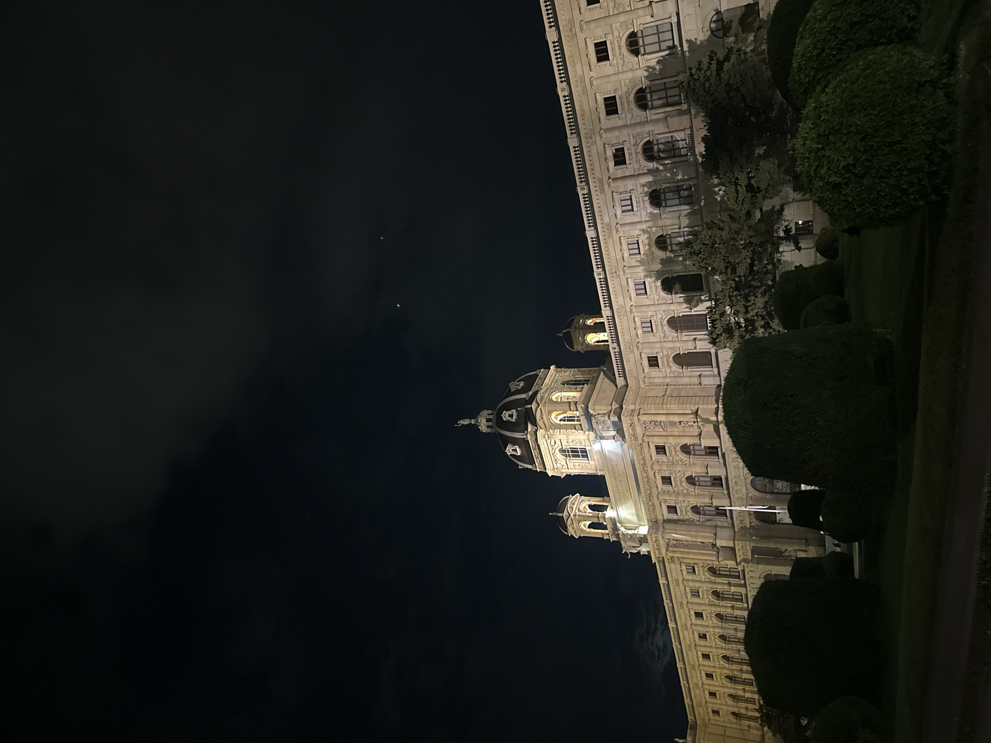 The image shows the exterior of the Natural History Museum (Naturhistorisches Museum) in Vienna, Austria, at night. The imposing, ornate building is illuminated by artificial lights, highlighting its Baroque architecture, including a large central dome and numerous windows. In the foreground, neatly trimmed hedges and trees form part of a garden or park setting, casting shadows on the well-maintained lawn. The sky above is dark with some clouds, and a few faint lights, possibly stars or distant aircraft, are visible. There are no people or vehicles visible in the frame.