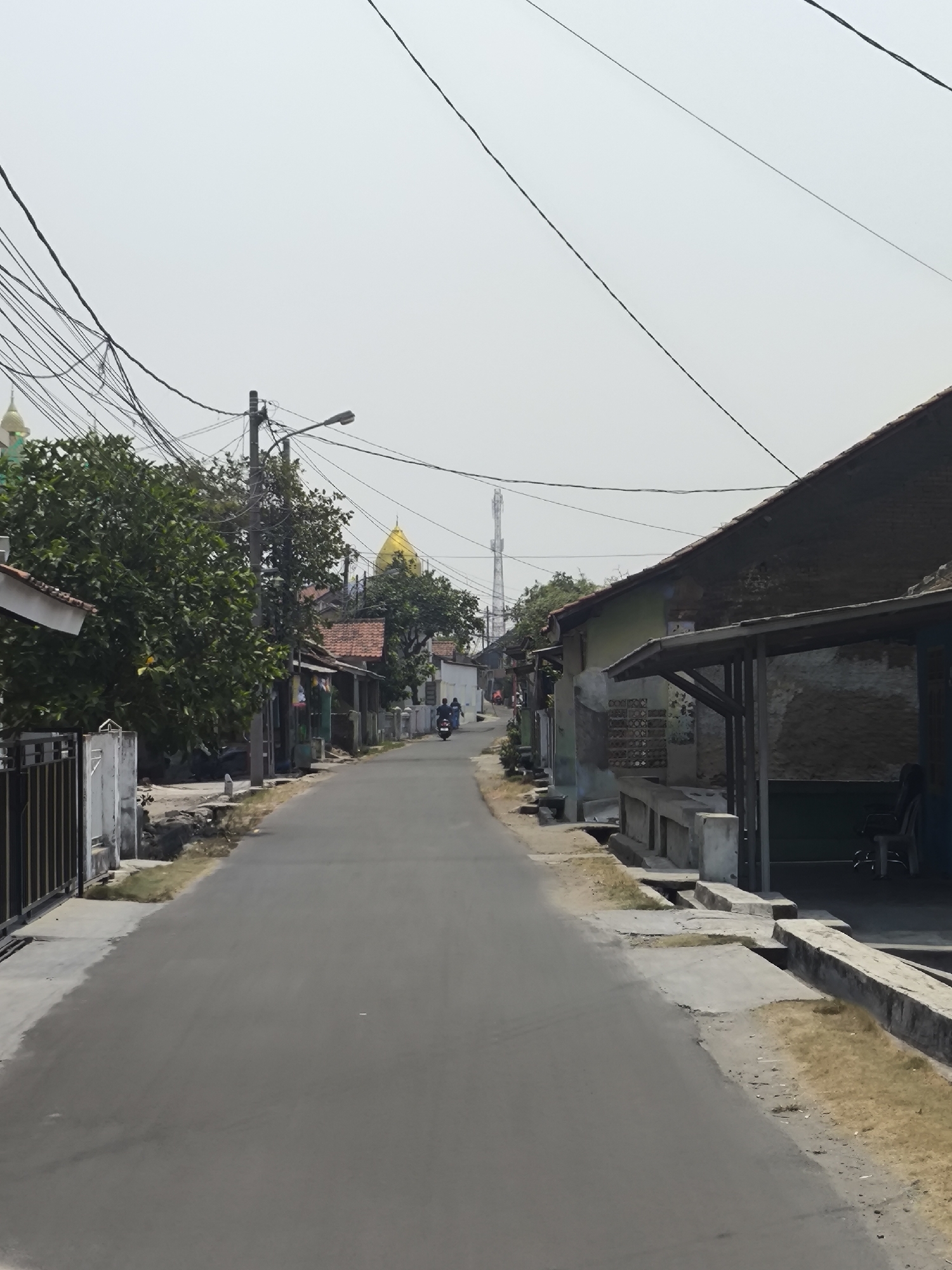 This image shows a street scene in Cilegon, Indonesia, under a bright, overcast sky. The street is paved and appears to be a residential or small commercial area. On the left side of the street, there are fences and buildings with trees. A notable structure with a green dome is visible further down the street on the left, and a golden dome is visible on the right, suggesting religious buildings are present in the vicinity. Power lines are strung across the sky, with a street lamp visible on the left side of the road. On the right side of the street, there are buildings with covered overhangs, and some concrete structures. A telecommunications tower can be seen in the distance. Two people are riding a motorcycle down the street. The environment appears to be a typical urban or semi-urban setting in Indonesia.