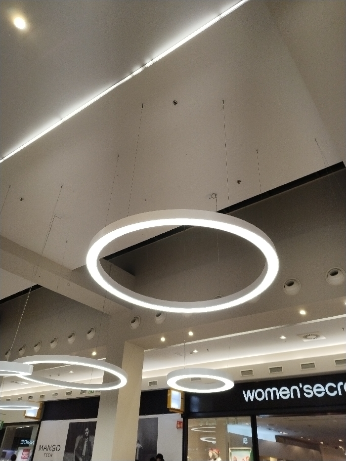 The image showcases the interior of a shopping center with multiple ring-shaped light fixtures suspended from the ceiling. The fixtures are uniformly illuminated, providing ambient lighting. Below, storefronts are visible, including "Mango Teen" and "Women'secret." The ceiling also features recessed lighting and a linear light strip along the top. The architecture is modern, characterized by clean lines and a neutral color palette.