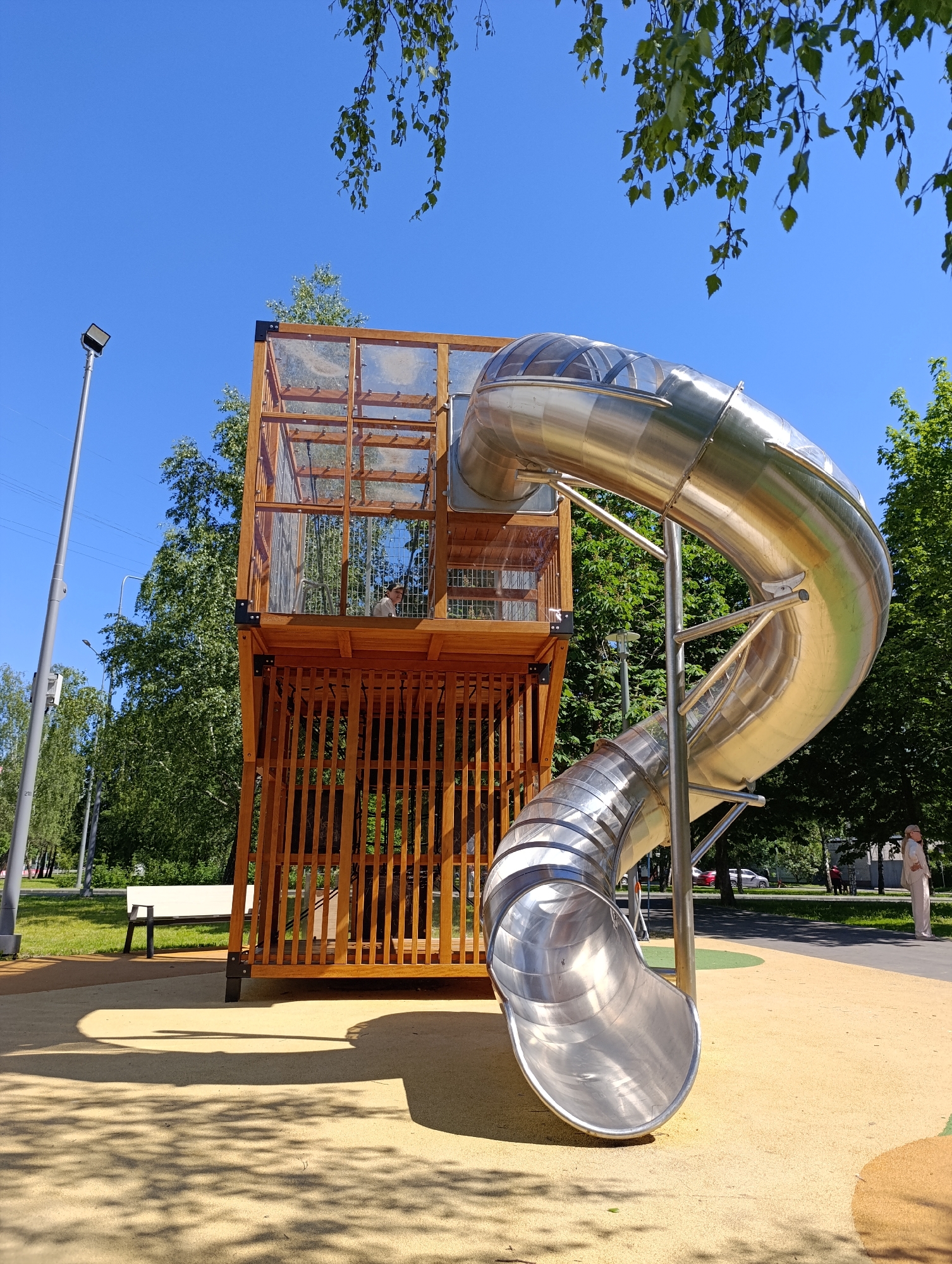 This image captures a modern public playground structure on a clear, sunny day in Moscow, Russia. The primary subject is a multi-story wooden tower featuring a large, curving, enclosed stainless steel slide. The lower portion of the wooden tower consists of vertical wooden slats, while the upper level is enclosed with transparent panels, providing visibility. One person, wearing a light-colored top, is partially visible through the upper transparent panels of the tower, looking towards the right side of the frame. The polished stainless steel slide spirals downward, supported by metal posts, and terminates on a light yellow/tan colored rubber safety surface, which covers the playground area. This surface exhibits some irregular darker orange/brown patches.
Strong sunlight illuminates the scene, casting distinct shadows from the playground structure and slide onto the ground, along with dappled shadows from overhead trees. In the background, numerous lush green deciduous trees indicate a park setting. A bright blue sky occupies the upper frame, with branches of a tree with light green leaves visible in the top corners. On the far left, a tall, gray street lamp with a rectangular light fixture stands upright. A white park bench is visible on a grassy patch behind the playground structure. On the far right, a person in light-colored attire, including a hat, stands near the edge of the safety surface, adjacent to a darker paved path or road where several vehicles are partially visible.