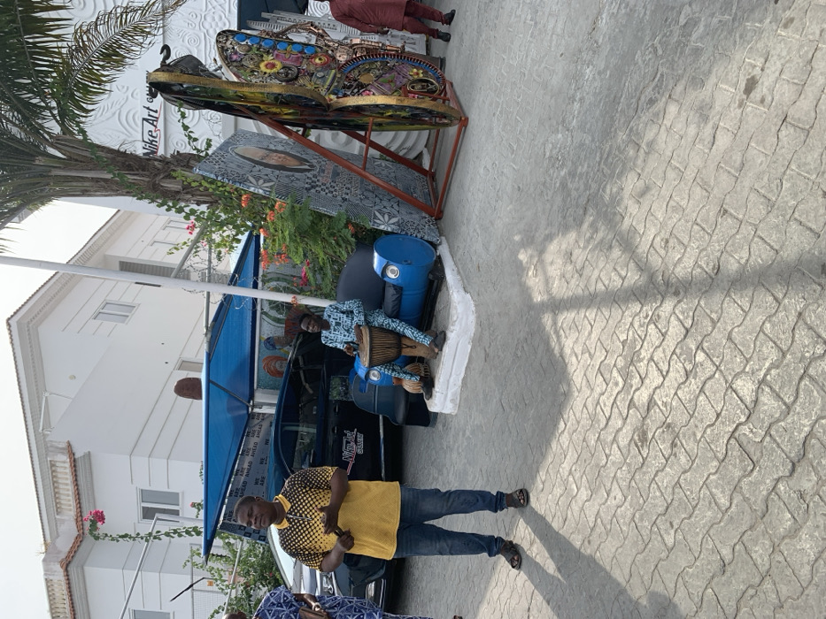 The scene depicts an outdoor paved area in front of the Nike Art Gallery in Ikate, Lagos, Nigeria, under natural daylight conditions.
In the foreground, a light grey interlocking paver surface extends across the ground, featuring distinct shadows from subjects and overhead structures.
On the left, a male subject stands, facing right, wearing a yellow and black patterned polo shirt, blue denim jeans, and dark sandals. His hands are clasped at waist level, appearing to hold a small, dark object.
To his right, a dark-colored sedan, possibly black, is parked beneath a blue fabric shade canopy. Text "WE ARE AHEAD" and "Nike Art Gallery" are visible on or near the vehicle.
Further right, a male subject is seated on a custom-made blue sofa, constructed from repurposed metal barrels, with a dark cushion. This subject wears a blue and white patterned long-sleeved shirt and matching trousers. He is actively playing a djembe drum positioned between his legs.
Behind the seated drummer, two large, vertical art installations are displayed. The left piece features a stylized face and geometric patterns in shades of grey and black. The right piece is a multi-colored, complex sculpture resembling an abstract butterfly or mechanical insect, adorned with various materials and textures.
The background features two large white buildings. The structure on the left has multiple windows and a balcony with pink bougainvillea vines. The building on the right, identified by a sign as "Nike Art Gallery," exhibits textured, wave-like patterns on its facade. A tall palm tree is visible on the far right, alongside various green foliage and plants with orange-red flowers.
Additional subjects are partially visible: one on the far left, wearing patterned attire, and another on the far right, wearing a red/maroon outfit. No direct interactions between the visible subjects are clearly depicted.