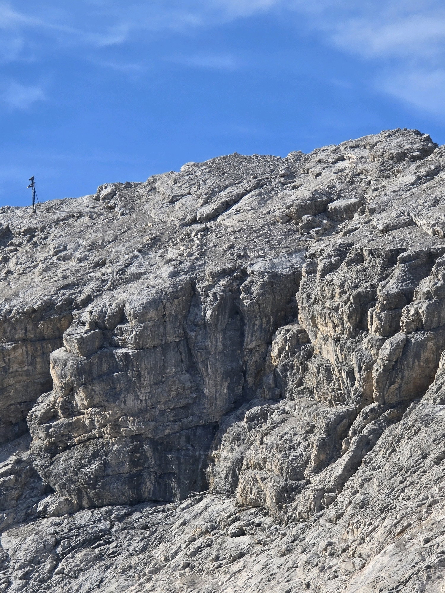 A steep, rocky mountainside with a bright blue sky above. A dark, narrow pole with an object at the top stands on the crest of the mountain in the upper left quadrant of the image. The rock face is textured with numerous cracks, ledges, and outcroppings, giving it a rugged appearance. The lighting suggests it is daytime, with sunlight casting shadows that highlight the terrain's contours. The overall scene depicts a natural, mountainous landscape, likely in an alpine region. The image does not show the city of Garmisch-Partenkirchen, Germany.
