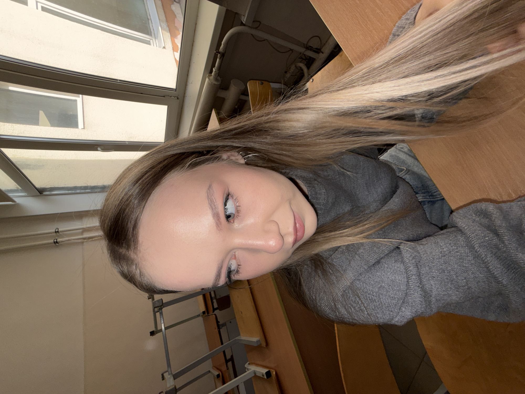 A young woman with light blonde hair, wearing a grey long-sleeved shirt, is photographed indoors. She is seated at a wooden desk with attached seats typical of a classroom or lecture hall setting. A window with multiple panes is visible behind her, suggesting an institutional building. The interior scene includes light-colored walls, and other desks are visible. Based on the classroom design and the presence of radiator pipes running along the wall near the window, the scene is likely a university or school within Chaikovsky, Russia.