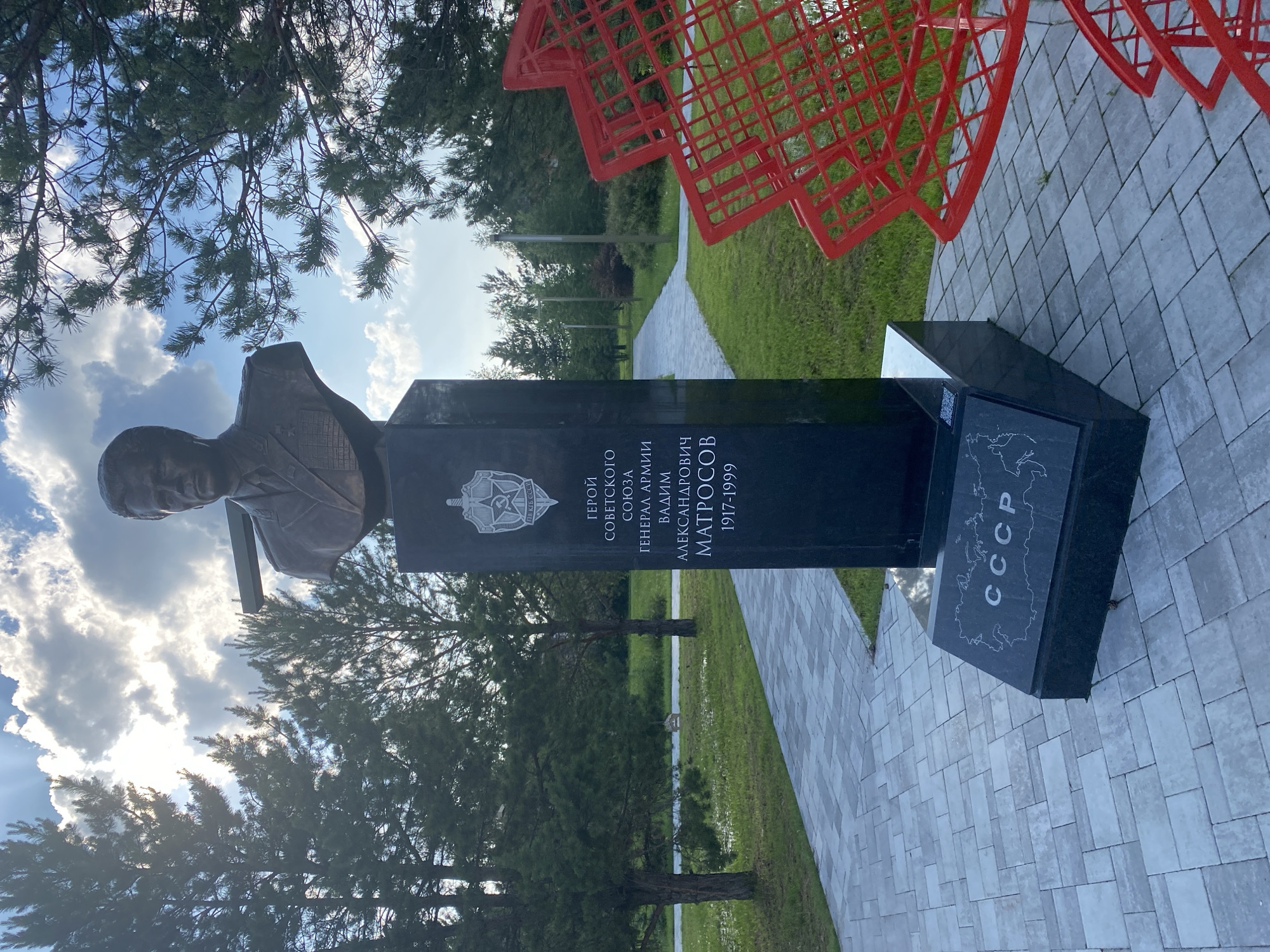 A bronze bust of General of the Army Vadim Aleksandrovich Matrosov (1917-1999) stands on a tall, black polished granite pedestal in the Square of Border Guards (Сквер Пограничников) in Tyumen, Russia. Matrosov is depicted in a military uniform, wearing medals and epaulets. The pedestal's upper section features the emblem of the KGB with "КГБ СССР" inscribed, followed by the text in Cyrillic: "ГЕРОЙ СОВЕТСКОГО СОЮЗА ГЕНЕРАЛ АРМИИ ВАДИМ АЛЕКСАНДРОВИЧ МАТРОСОВ 1917-1999" (HERO OF THE SOVIET UNION GENERAL OF THE ARMY VADIM ALEKSANDROVICH MATROSOV 1917-1999). The base of the monument is adorned with an etched map of the Soviet Union and the inscription "СССР." The monument is situated on light grey interlocking paver stones, which form a walkway leading into a park-like setting. The surrounding area features manicured green grass and numerous mature coniferous trees, primarily pines, with sunlight filtering through their branches, creating visible rays. A partially visible, large, red, abstract, grid-like sculpture is located to the right of the monument. The sky above is blue with scattered white clouds, and the sun is bright, indicating daylight conditions.