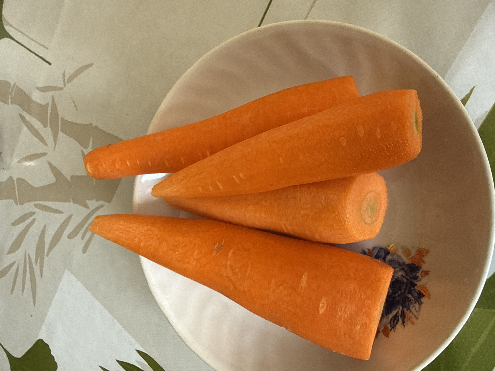 Four peeled carrots, cut into conical shapes with the pointed ends facing up, are arranged in a white bowl on a tablecloth. The tablecloth has a light cream background with a pattern of grey bamboo stalks and green leaves. The bowl has a floral design on its interior. The scene is indoors. The location is not specified.