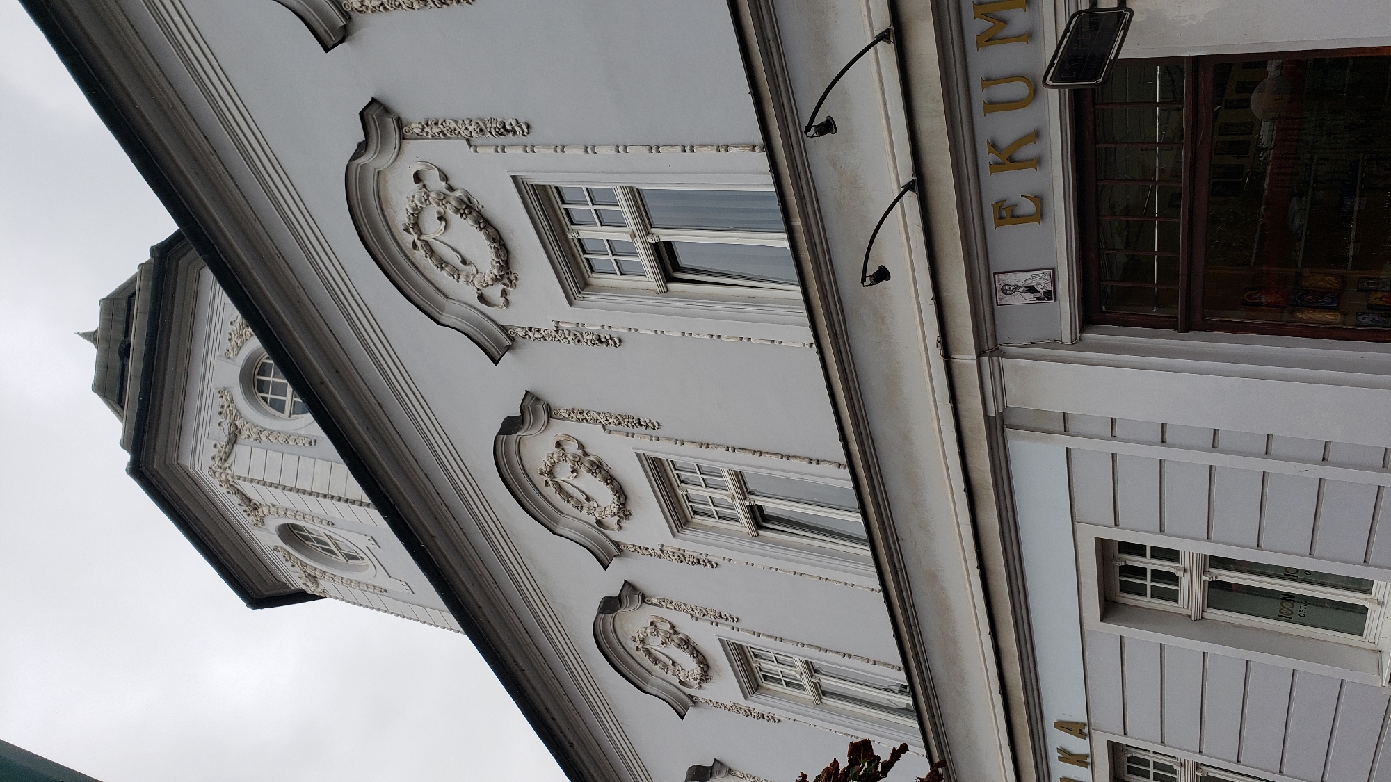 This is a low-angle exterior shot of a building in Prešov, Slovakia. The building features pale gray walls with ornate white decorative elements around its windows and upper facade. The windows on the upper floor are arched and feature decorative garlands and vertical line ornamentation. The building has a tower-like structure at the top with a spire. Below the decorative upper facade is a simpler section with horizontal paneling and more windows, some with signs indicating "ICON OPTIC." Further to the right, there is a storefront with the golden letters "EKUM" visible above a dark-framed window. A small framed image of a person in a religious robe is mounted on the wall above the storefront. The sky is overcast.