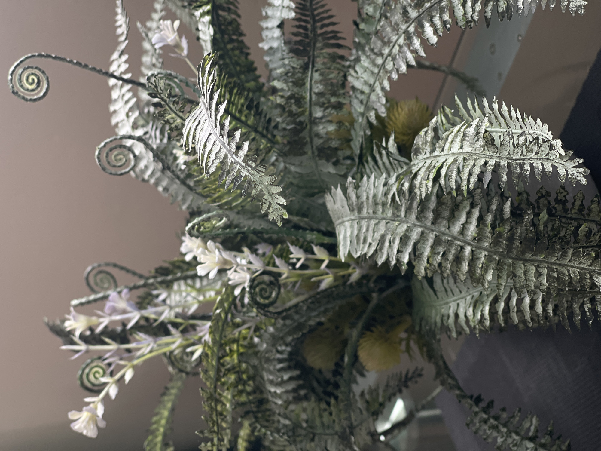 The image shows a close-up of a floral arrangement, primarily composed of artificial ferns and small white flowers. The ferns exhibit a distressed or aged appearance with a dusty, silvery-green hue. Some fern fronds are curled in a spiral shape, characteristic of fiddleheads. Interspersed among the ferns are delicate stems with small white blossoms and a few yellow, spherical elements that could be artificial berries or seed pods. The arrangement is placed in a container, the edge of which is partially visible at the bottom of the frame. The background is a plain, muted color, likely a wall. The lighting suggests an indoor setting, with a soft, diffused light source. There are no people or animals visible in the image. The image's composition focuses on the textures and details of the artificial plants. There is no information in the image to indicate a location in Bolshoy Kamen, Russia.