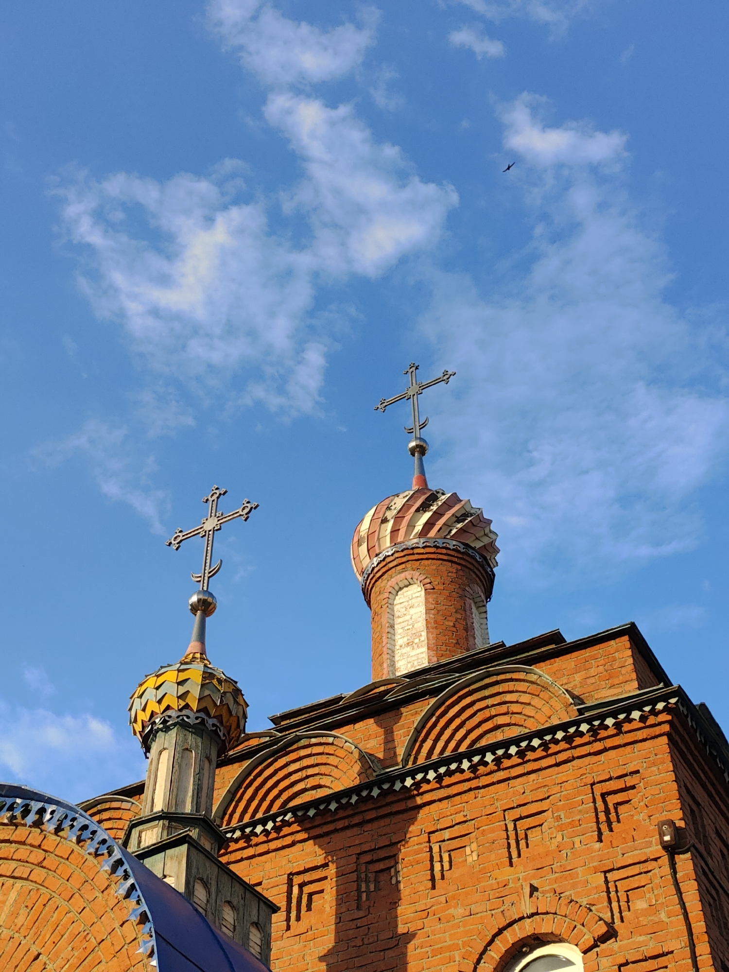 The image shows the domes and brickwork of an Orthodox church, possibly in Ufa, Russia, under a bright blue sky with scattered white clouds. Two crosses top the domes. The foreground features a large, elaborately decorated brick wall section of the church, with arched details and patterned brickwork. One dome is covered in colorful, scale-like tiles in yellow, blue, and gray, while the other is primarily red and white striped. The architectural style suggests a historical or traditional Orthodox church. A bird is visible in flight in the upper right portion of the sky.
