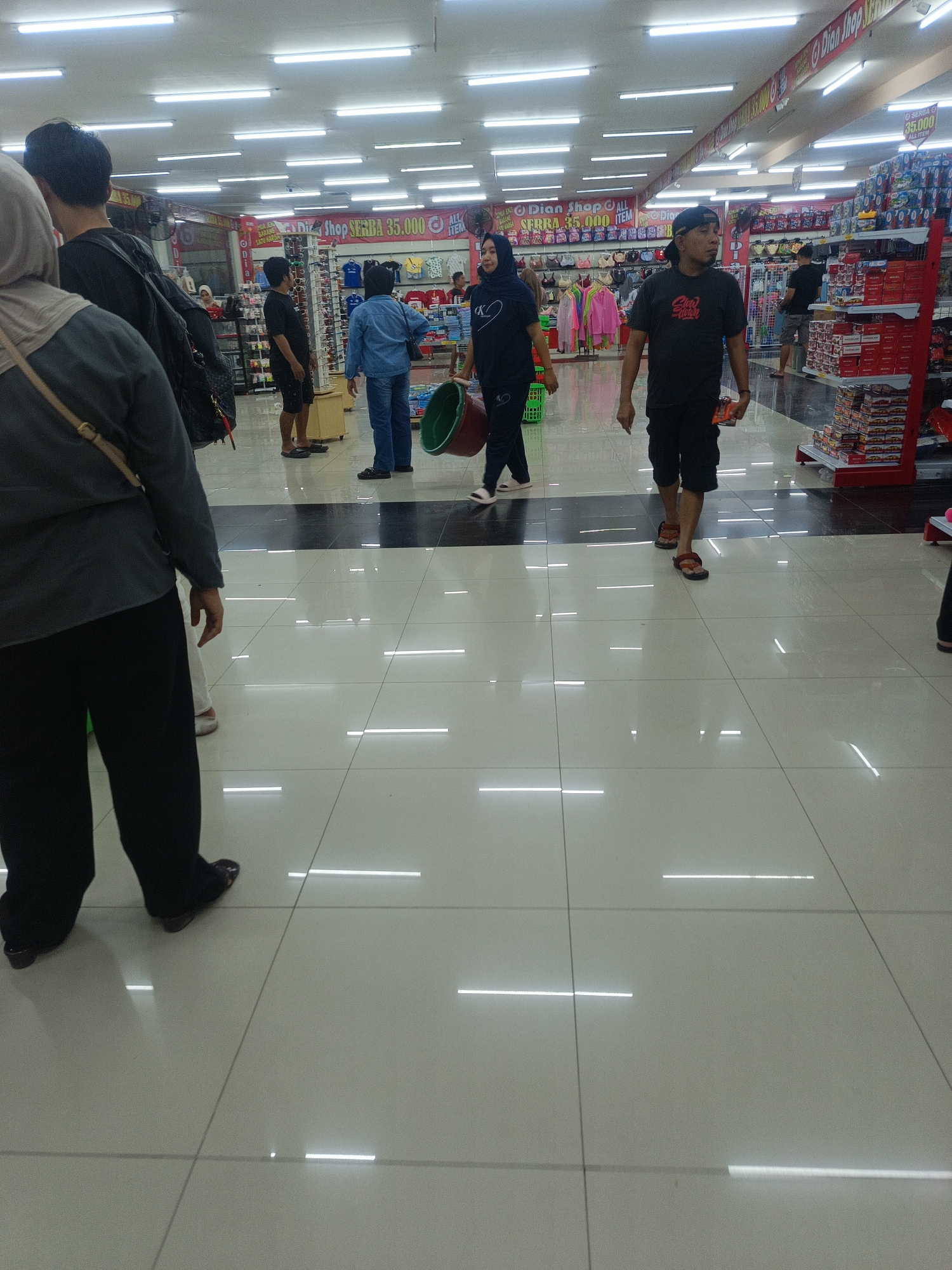 The image captures an interior scene within a commercial establishment, likely a department store or supermarket, in Limboto, Indonesia. The environment is well-lit with fluorescent tube lighting lining the ceiling. The floor is tiled with a combination of white and black squares.
Several individuals are present, walking and interacting within the store. In the foreground, on the left, a person is seen from the back, wearing a dark grey long-sleeved shirt and black pants. Another person to their right is also partially visible, wearing black pants and dark footwear.
Further into the store, a man wearing a black t-shirt, black shorts, and a black baseball cap is walking towards the viewer's right, holding a small item in his right hand. To his left, a woman in a black hijab and black pants is walking away from the viewer, carrying a green bucket in her right hand.
To the left of the woman with the bucket, a person in a blue shirt and jeans is standing, facing away from the camera. Behind them, a man in a black t-shirt and dark shorts is walking towards the left.
Various merchandise displays are visible in the background, including racks of clothing, shelves stocked with products, and signage. Some of the signage appears to be in Indonesian. The overall impression is that of a bustling retail environment.