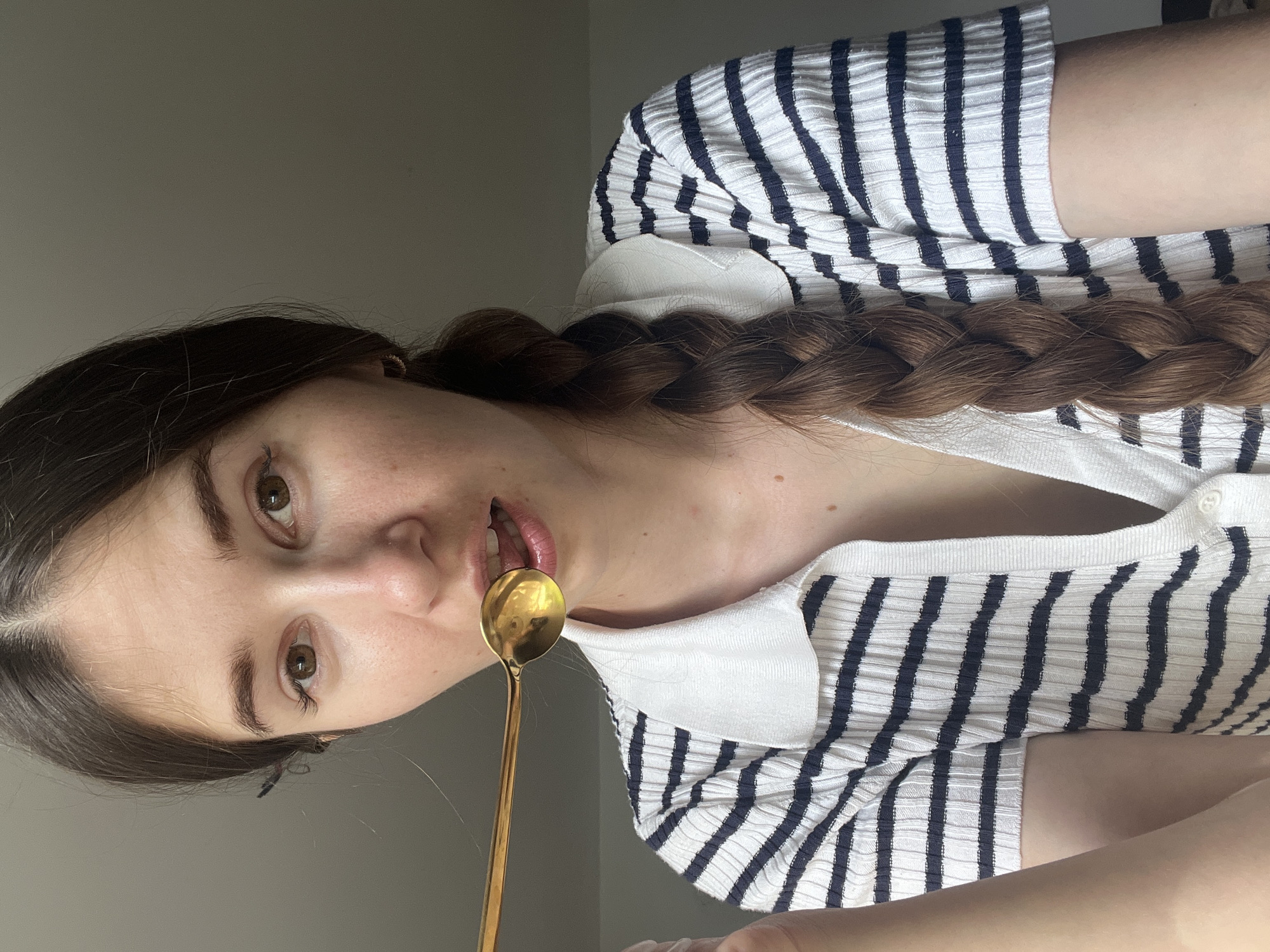 A young Caucasian woman with long, dark brown hair in a braid and brown eyes is shown from the chest up. She is wearing a white and navy striped short-sleeved shirt with a white collar and buttons down the front. The woman is holding a long-handled gold spoon to her mouth, with the spoon tip touching her lips. She appears to be looking at the camera. The background is a plain gray wall.