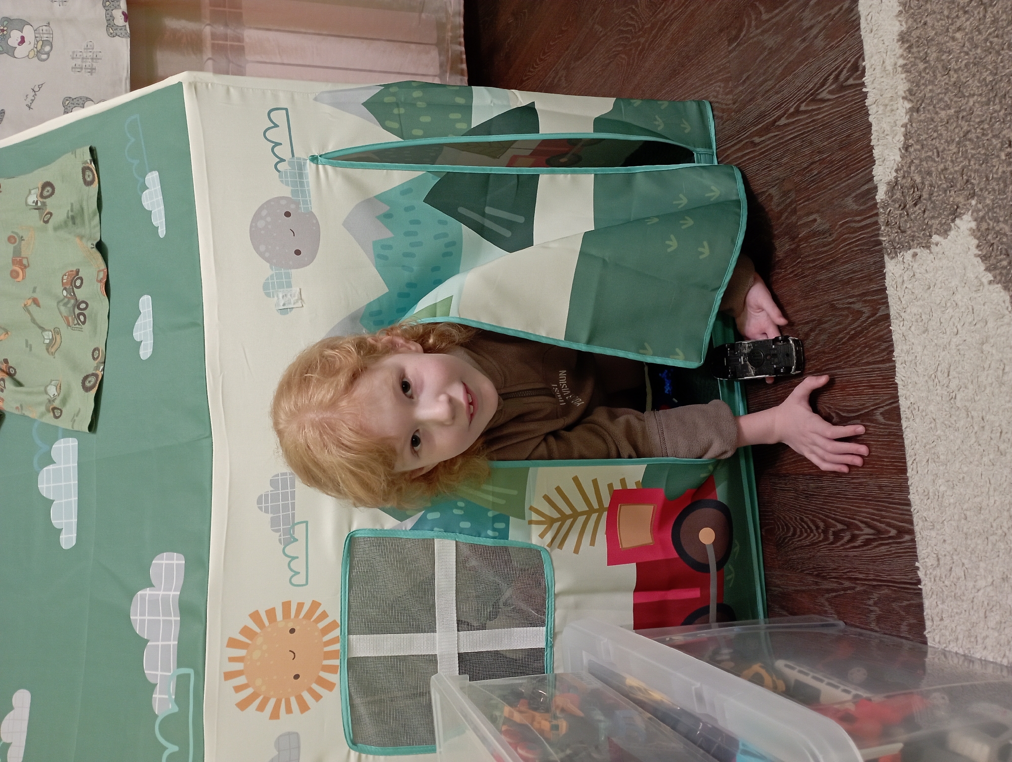 A young child with ginger hair and fair skin is peeking out from inside a play tent. The child is wearing a brown hooded sweatshirt. The tent has a green and cream colored exterior decorated with clouds, a sun, a moon, and mountains. A small black toy car is on the floor in front of the child. The play tent is situated on a wooden floor next to a clear plastic storage container filled with toys and a section of a cream-colored rug. The background shows a pink radiator and curtains. The image appears to be taken indoors in a residential setting, likely a child's play area.