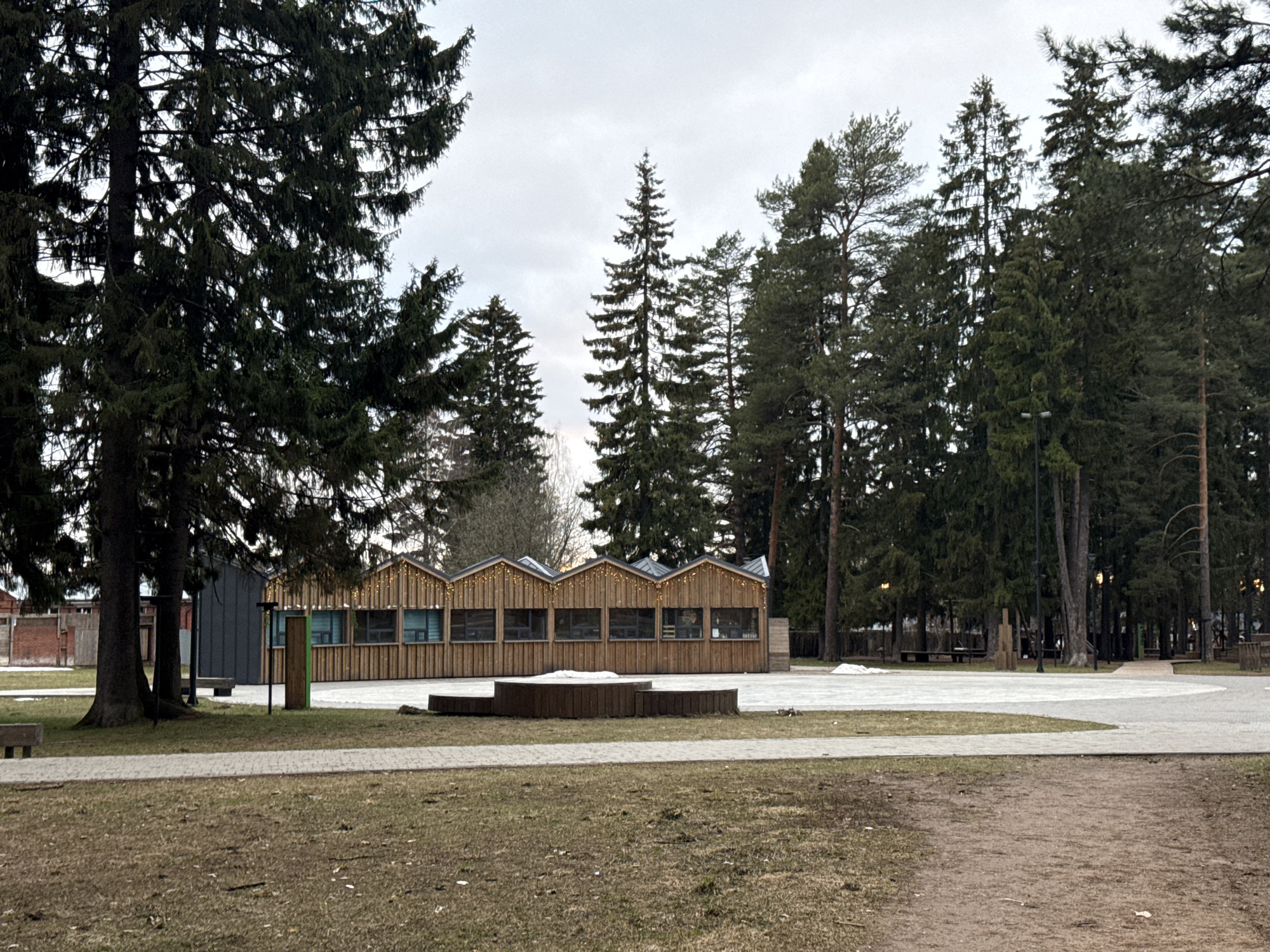 This image captures a quiet outdoor scene, likely a park or recreational area in Votkinsk, Russia, under an overcast sky. The ground exhibits patches of dry, light-brown grass and dirt, intermingled with light grey paved pathways and remnants of snow, suggesting late winter or early spring.
In the mid-ground, a prominent, long, low wooden building features a series of distinctive gabled roofs. Small, warm-toned decorative lights adorn the eaves of this structure. Its facade is constructed from vertical wooden planks, and it is punctuated by several rectangular windows with light-colored frames. Directly in front of this building, a low, multi-tiered circular wooden structure, possibly a planter or seating, sits on a paved area.
Numerous tall coniferous trees, appearing to be a mix of spruces and pines, define the landscape, framing the left and right sides of the image and extending into the background. Their dark green foliage contrasts with the pale, cloudy sky. On the far left, partially obscured by a large tree, is a darker, possibly utilitarian building, with a glimpse of a brick structure further behind it. A green trash bin is also visible near the large tree on the left.
The park is equipped with winding paved pathways. Further to the right, beyond the main wooden building, additional trees and various wooden park structures, including benches and playground elements, can be seen. Several streetlights are interspersed among the trees. The absence of people or any specific activity creates a tranquil and somewhat subdued atmosphere. No visible text is present in the image.