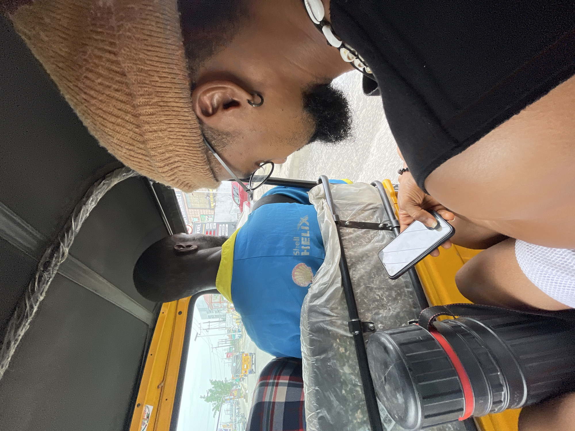 The image shows the interior of a yellow auto rickshaw in Ado, Nigeria. The rickshaw has a black roof and visible plastic sheeting. A passenger in a blue and yellow Shell Helix shirt sits in front. Behind, another passenger wears a tan hood and eyeglasses and is looking at a phone, held by a person with black shells on a necklace. The passenger next to them is wearing a white shirt. A black container with a red band sits on the floor. Street scenes of Ado, with buildings, advertisements, and other vehicles, are visible through the open sides of the rickshaw.